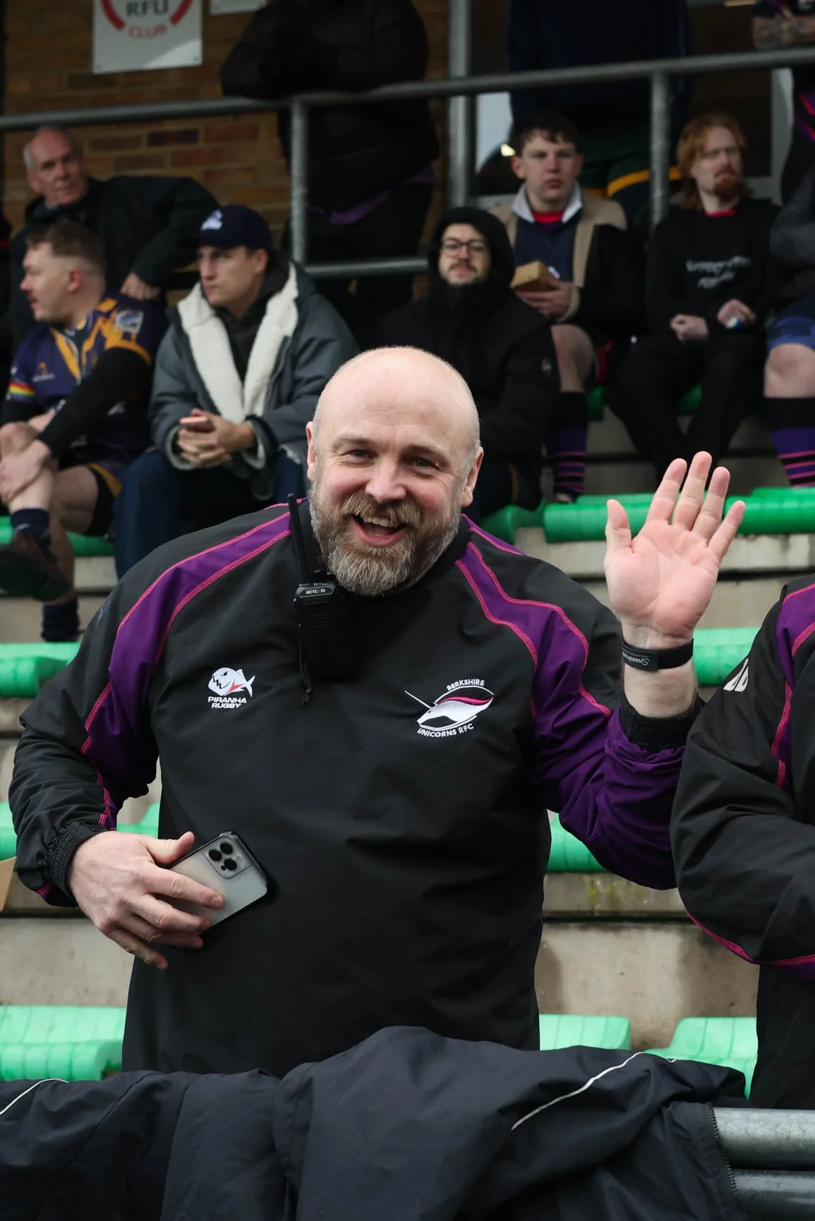 A smiling man in a black Berksih Unicorns rugby jacket, holding a phone and waving, standing among people in a stadium with green seating, other spectators in the background.