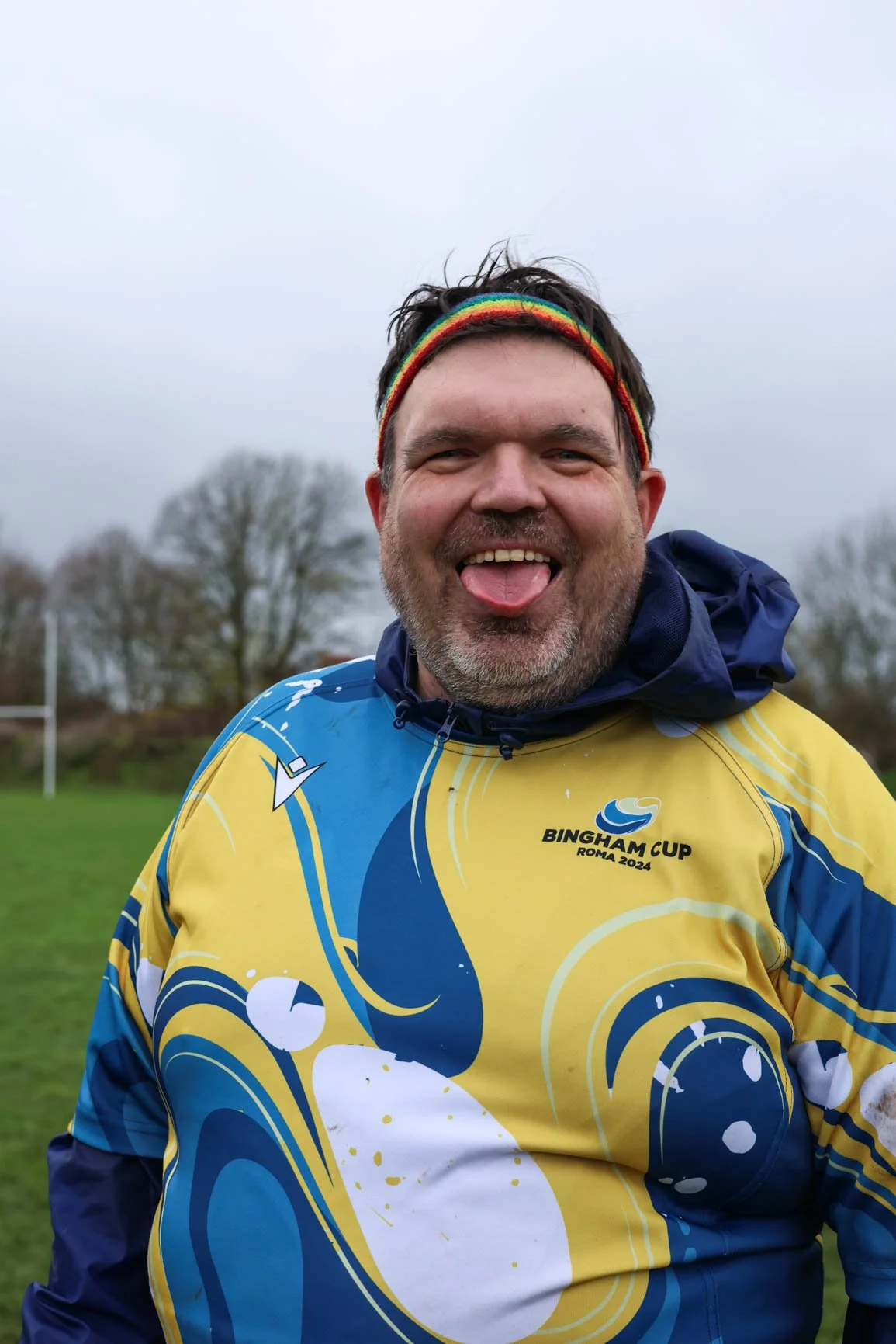 A man in a colorful sports jersey with a rainbow headband, smiling and sticking out his tongue outdoors on a cloudy day, with trees in the background.