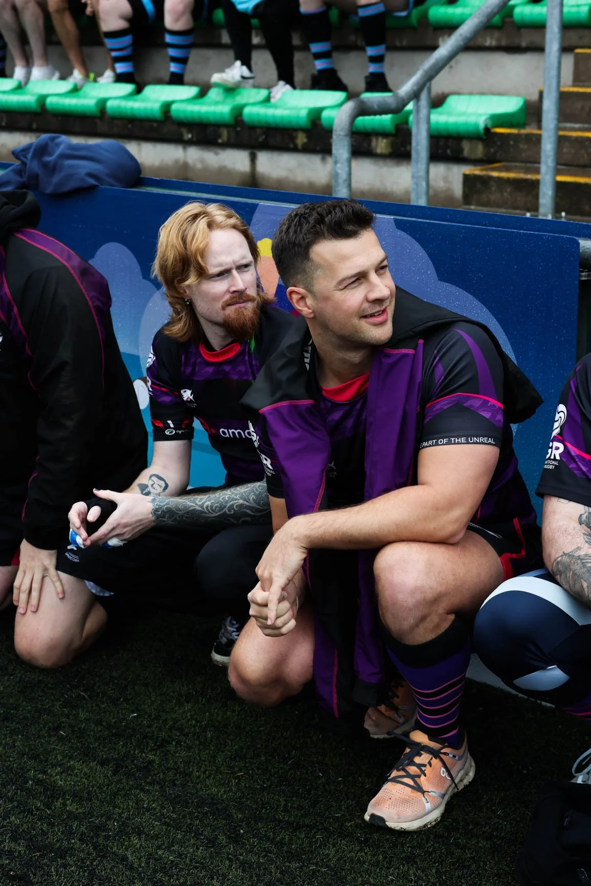 Two rugby players sitting on a bench, one with red hair and a beard, the other with dark hair, both wearing black and purple jerseys, with others in the background. The player with dark hair is smiling and looking away, while the red-haired player lo