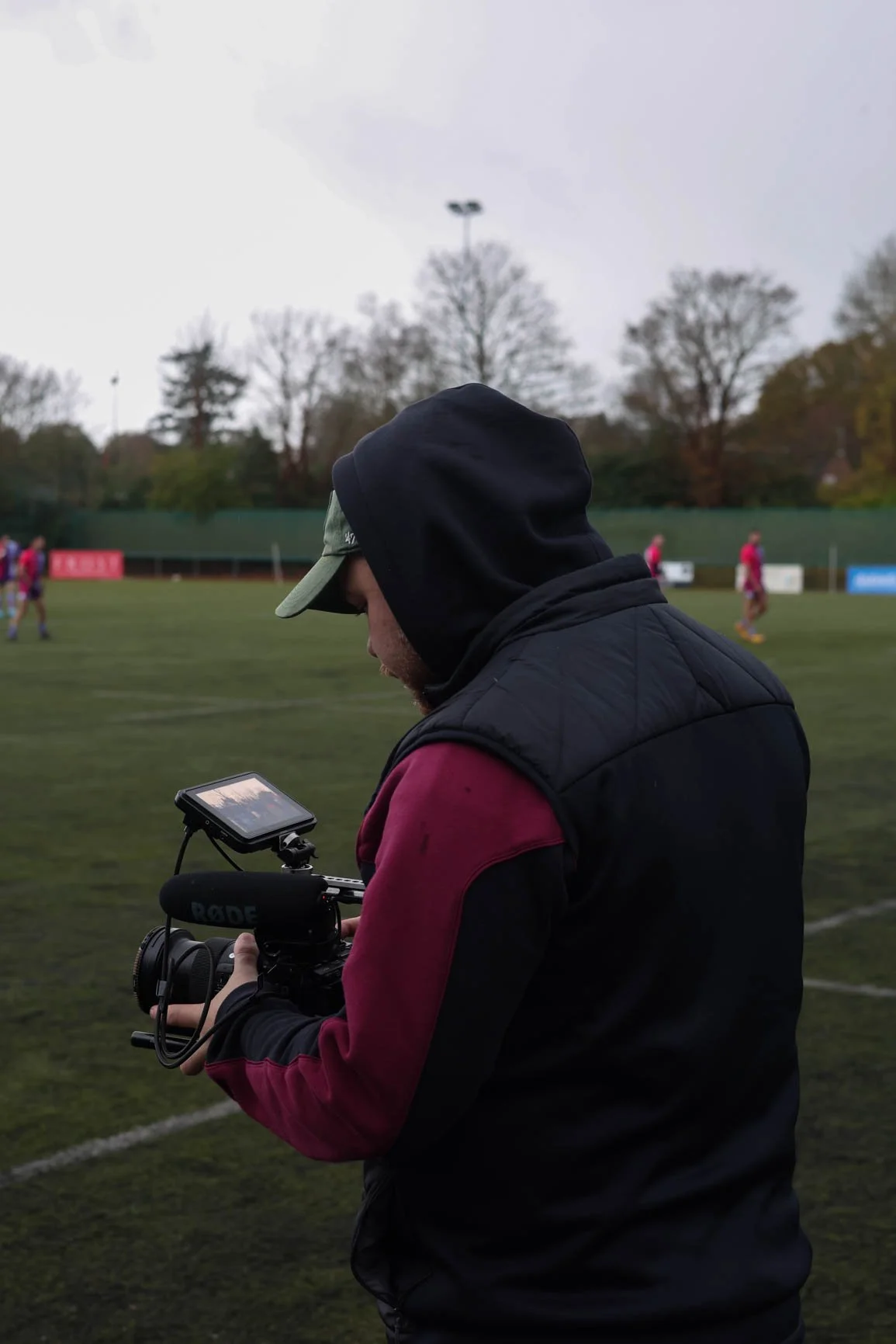 A person wearing a black hoodie and red jacket operating a camera with a monitor, on a sports field during a cloudy day, with players in red jerseys in the background.