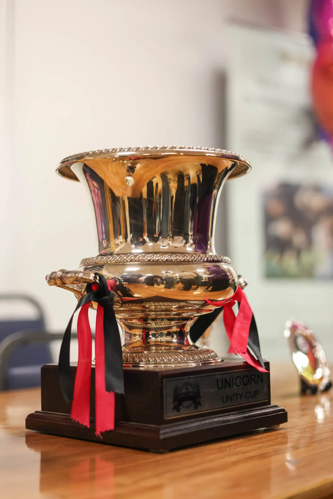A shiny gold trophy cup with red, black, and pink ribbons on a wooden table. The label on the trophy reads 'Unicorn Unity Cup.' In the background, there are blurred objects and a wall.