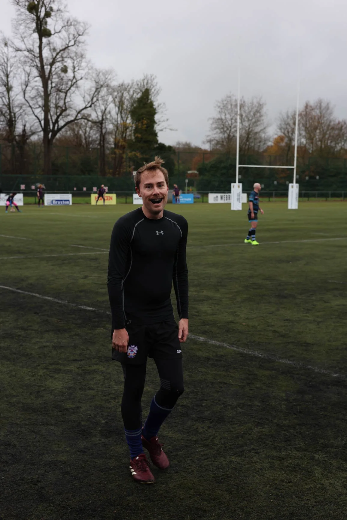 A man in black athletic clothing standing on a rugby field, smiling at the camera with rugby goalposts and other players in the background.