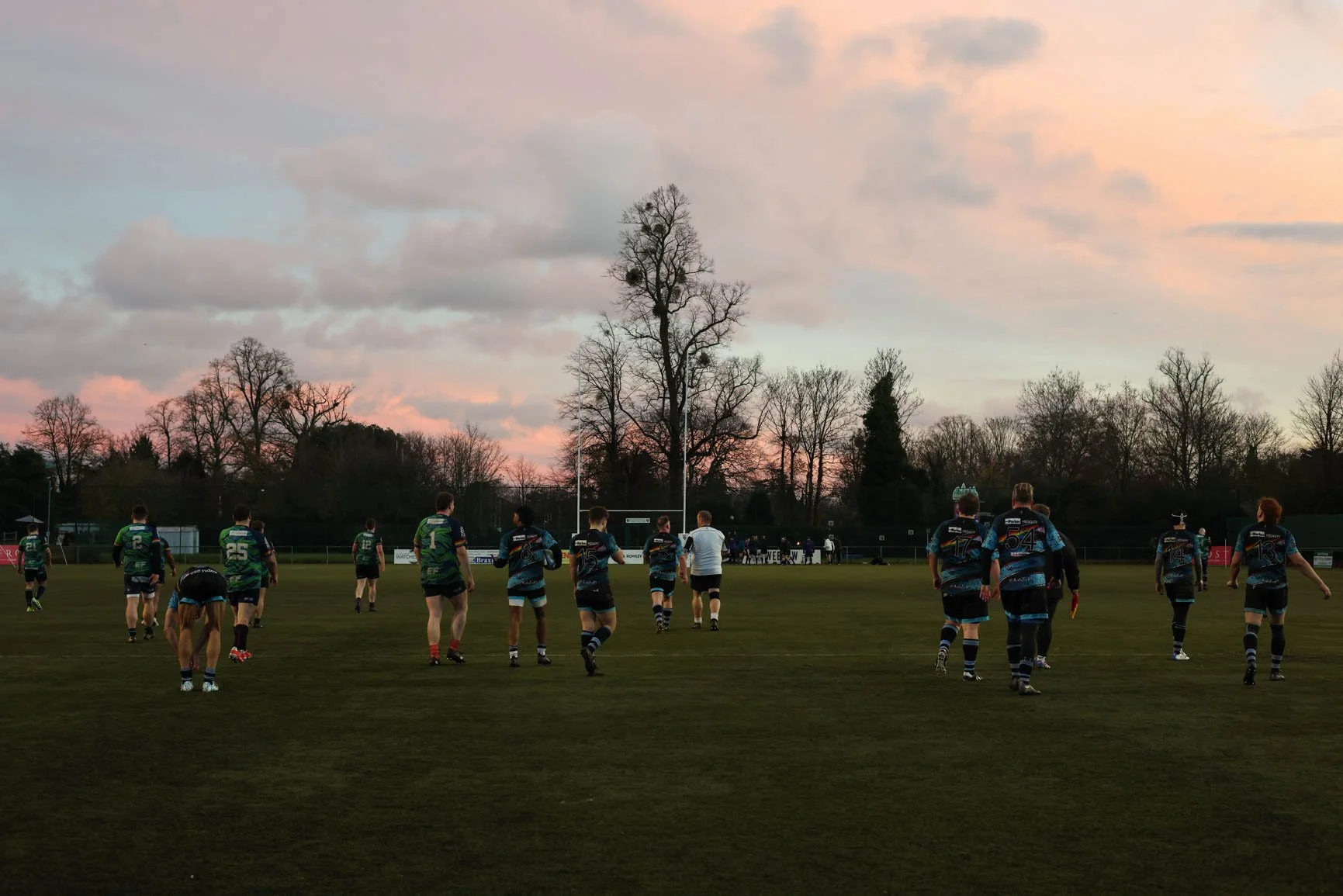 Rugby players walking onto the field during sunset, with tall trees and cloudy sky in the background.