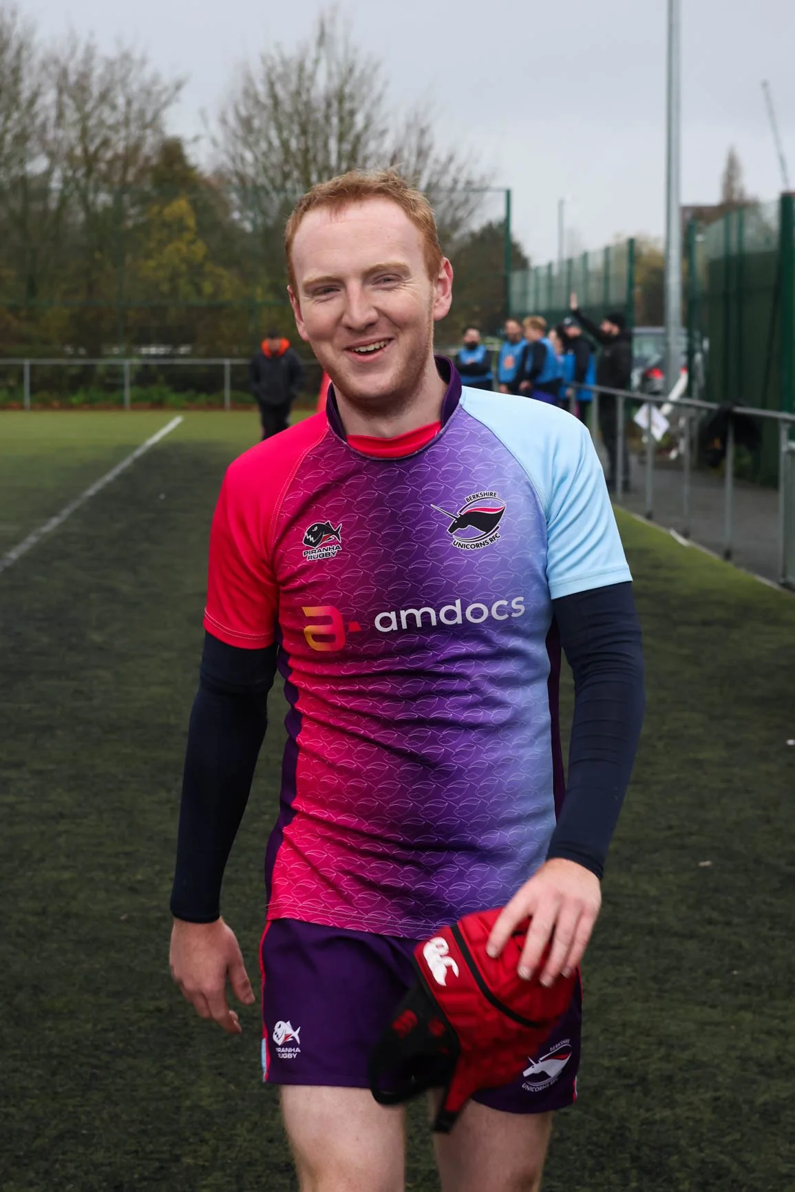 A young man in a colorful rugby jersey holding a red and black rugby helmet on a sports field, smiling at the camera with people and trees in the background.