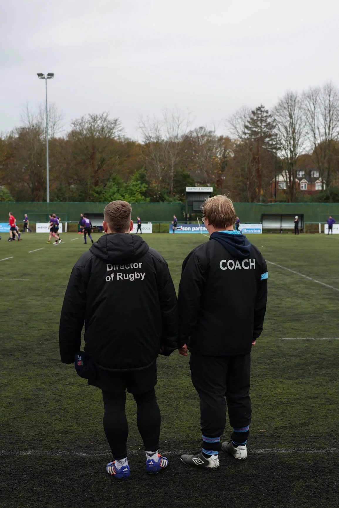 Two rugby coaches dressed in black jackets with one labeled "Director of Rugby" and the other "Coach" stand on the sideline of a rugby field watching players practice under an overcast sky, with trees and houses in the background.