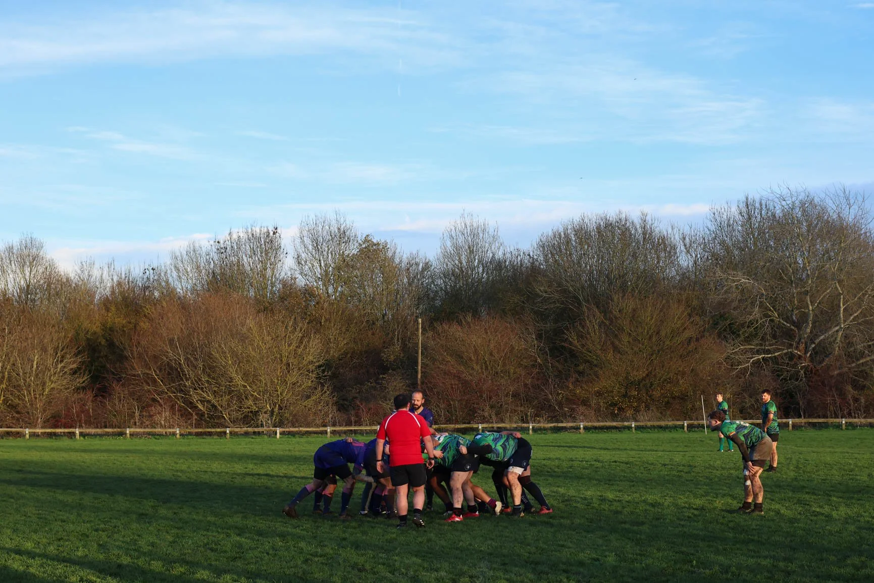 A rugby team in green uniforms huddles on a grassy field with players preparing for a scrum, while other players stand nearby, against a backdrop of leafless trees and a blue sky.