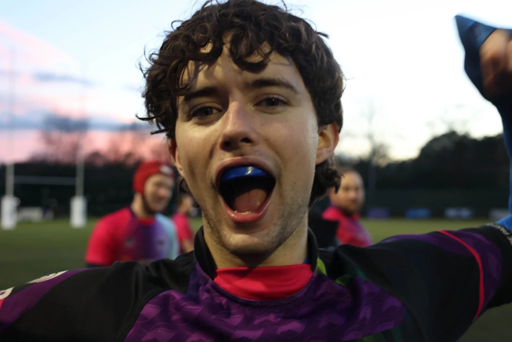 A young man on a sports field taking a selfie, with his mouth open and a blue mouthguard visible, and others blurred in the background during sunset.