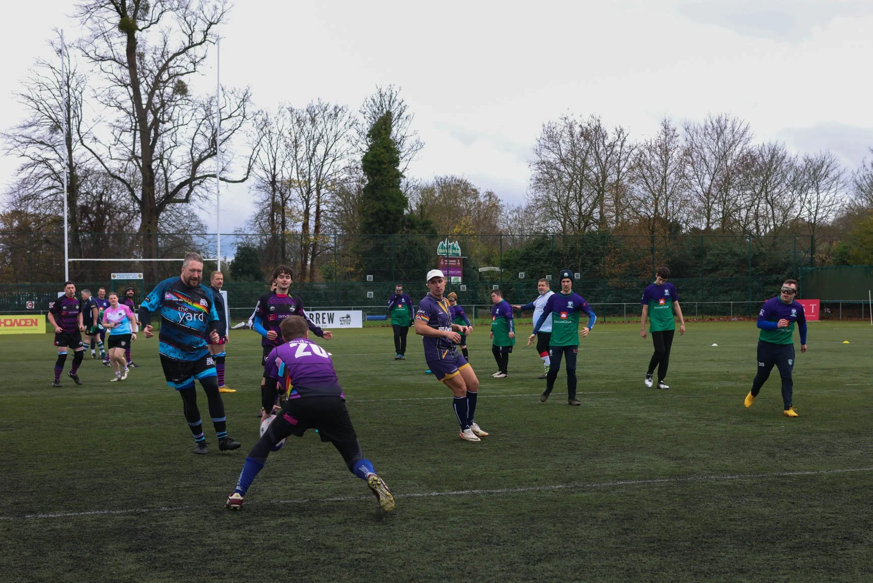 A group of people playing or practicing rugby on a grass field, wearing sports uniforms, with leafless trees and a cloudy sky in the background.
