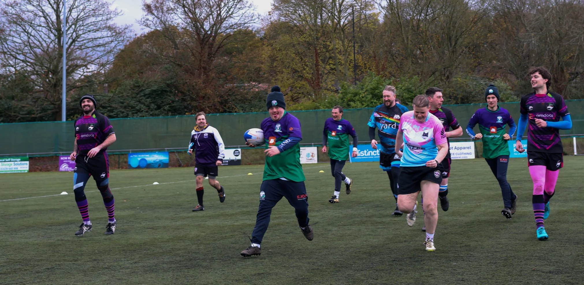 A group of rugby players, in colorful uniforms, running and smiling on a grassy field during a practice or game, with trees and advertising banners in the background.