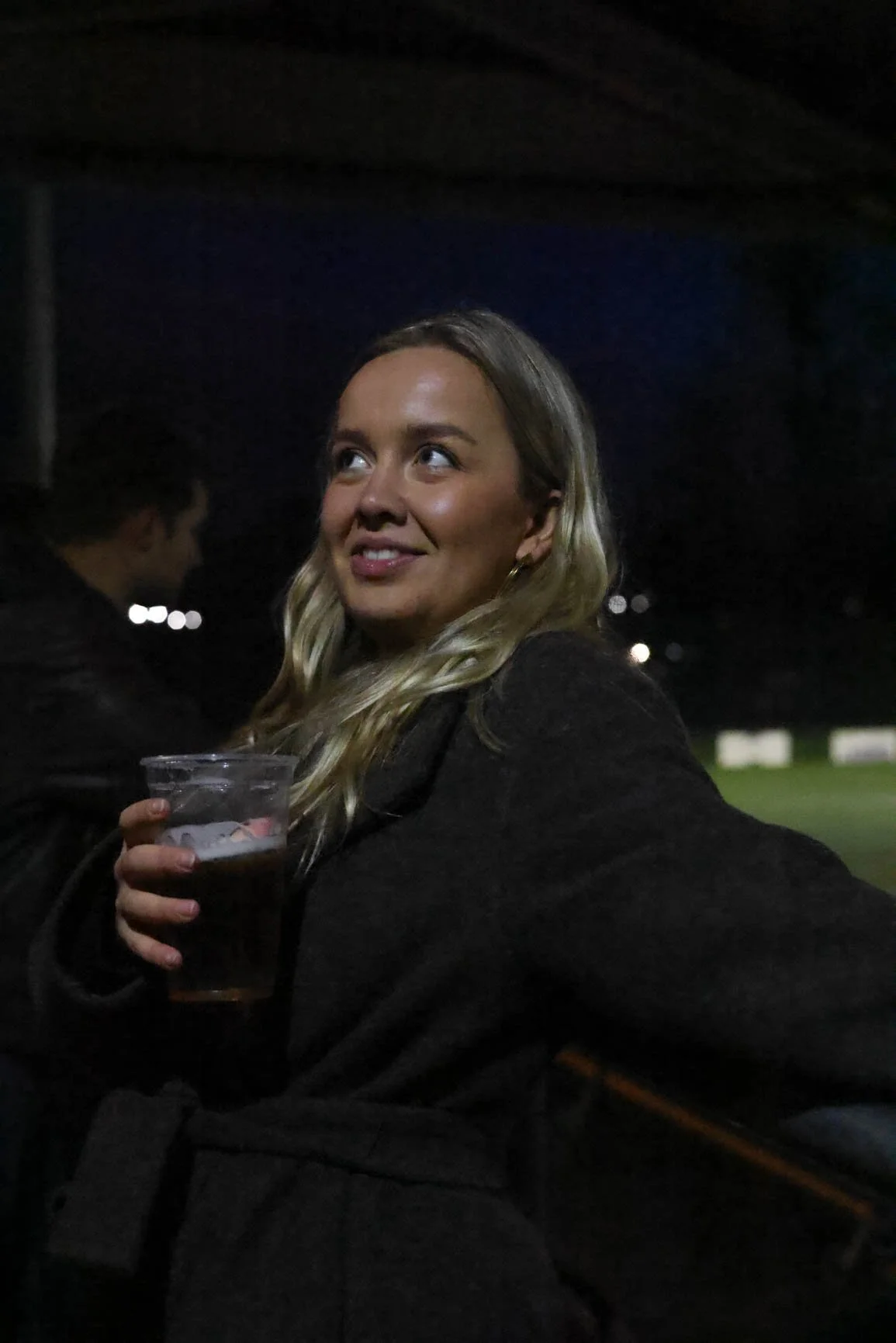 A woman with blonde hair smiling and holding a clear plastic cup with a dark beverage at night outdoors.