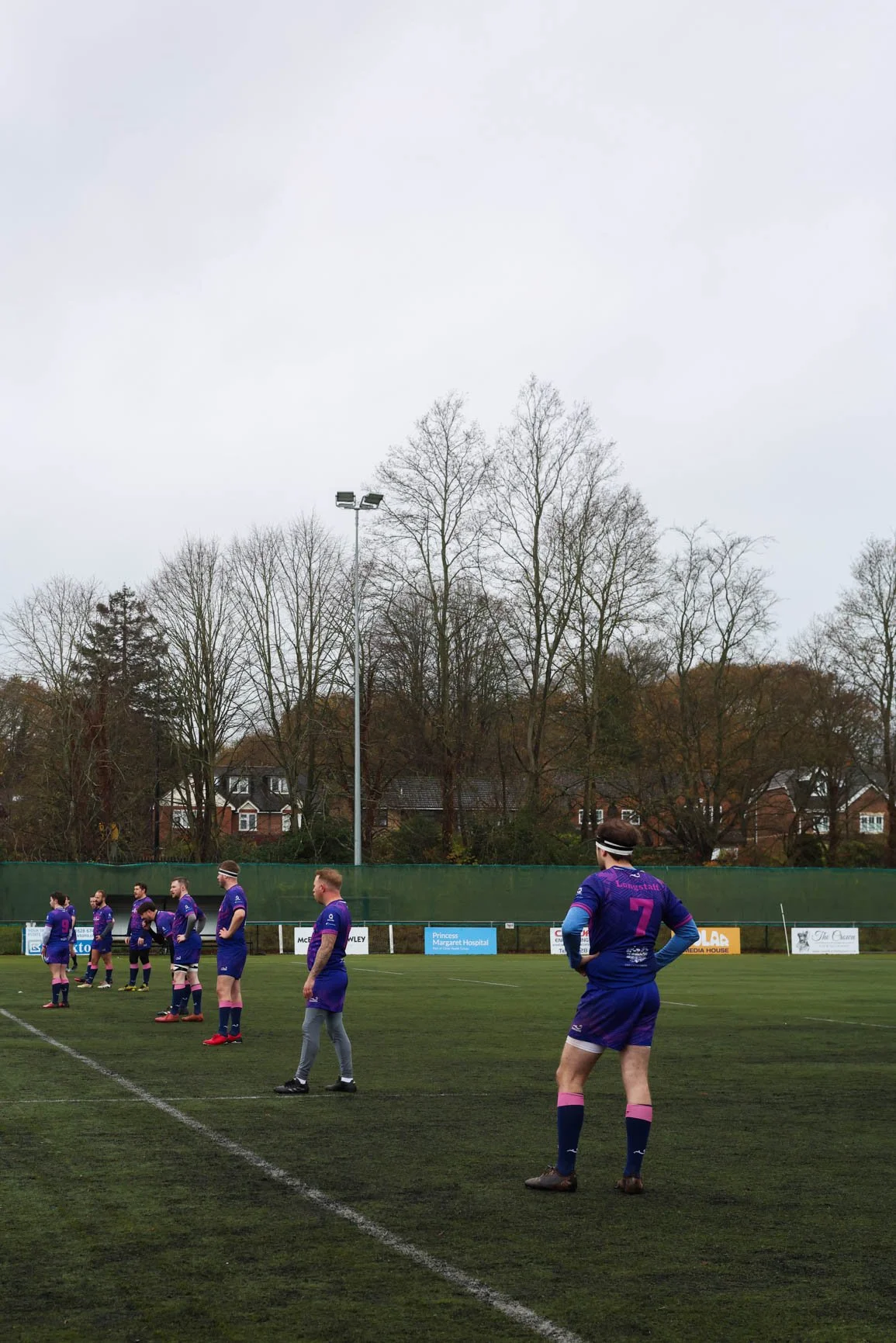 A group of rugby players in purple uniforms standing on a field during a game, with a few trees, buildings, and a cloudy sky in the background.