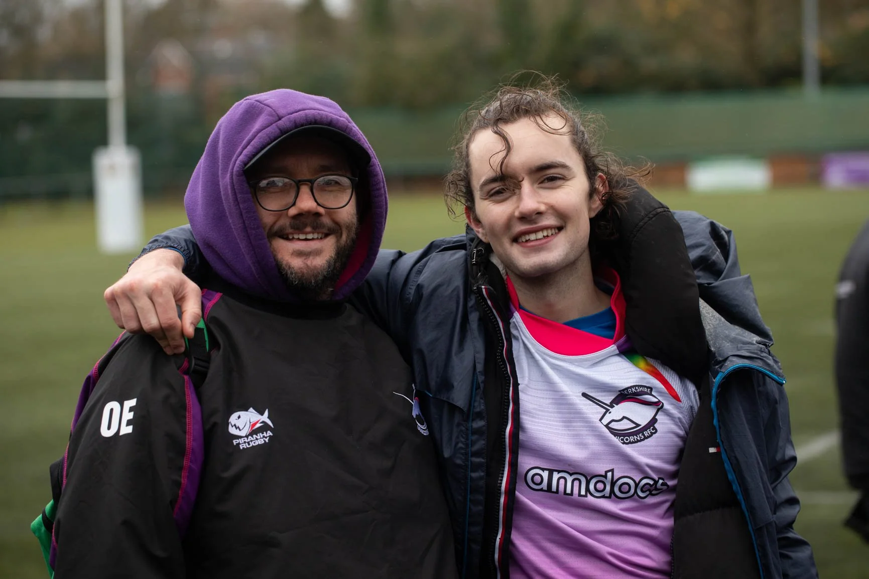 Two men are smiling and hugging outdoors on a grassy field, wearing sports jackets and rugby jerseys. One is wearing glasses and a purple hood, the other has curly hair and is wearing a rugby jersey.