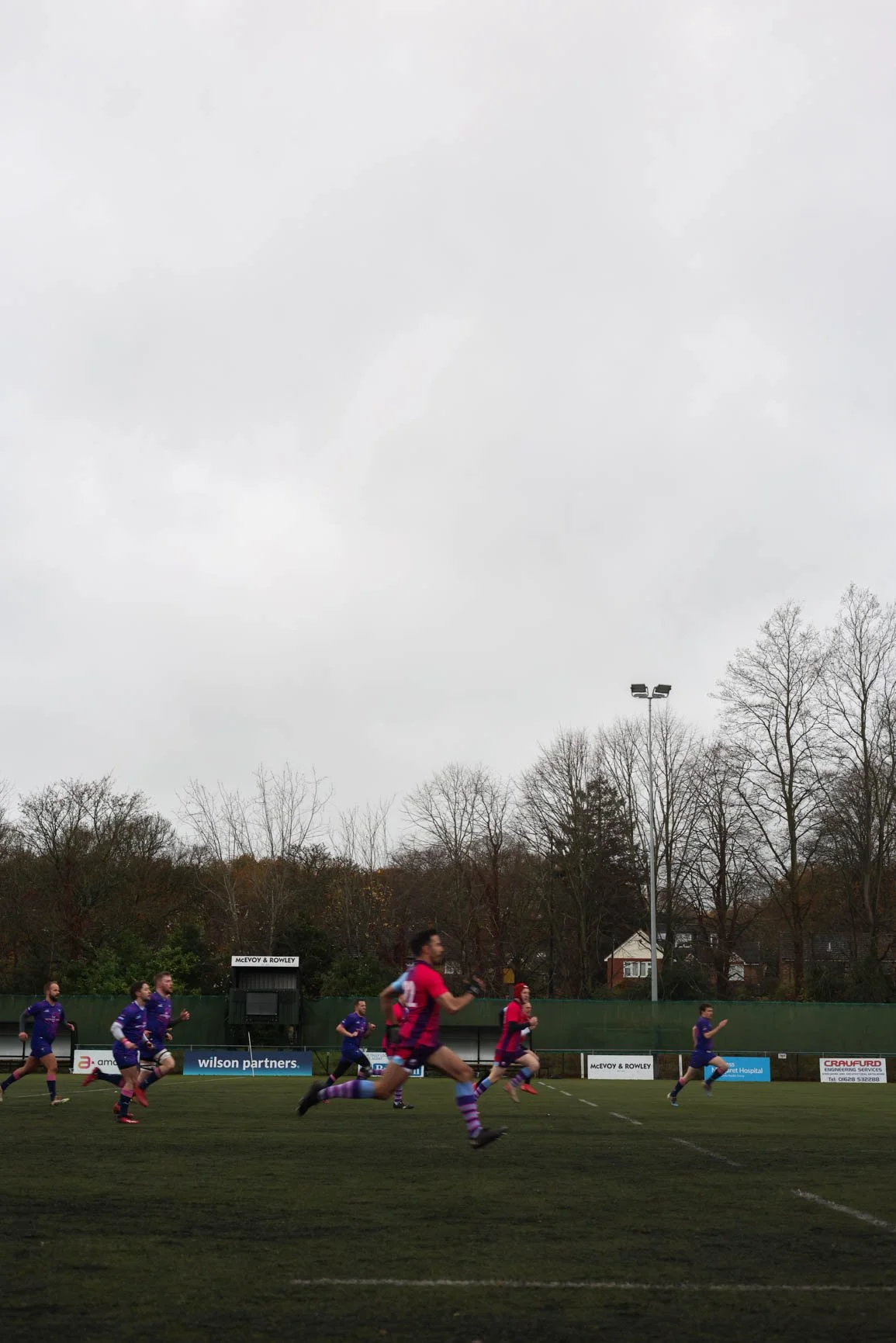 A group of soccer players in purple and red uniforms playing a match on a grassy field under an overcast sky, with leafless trees and some houses in the background.