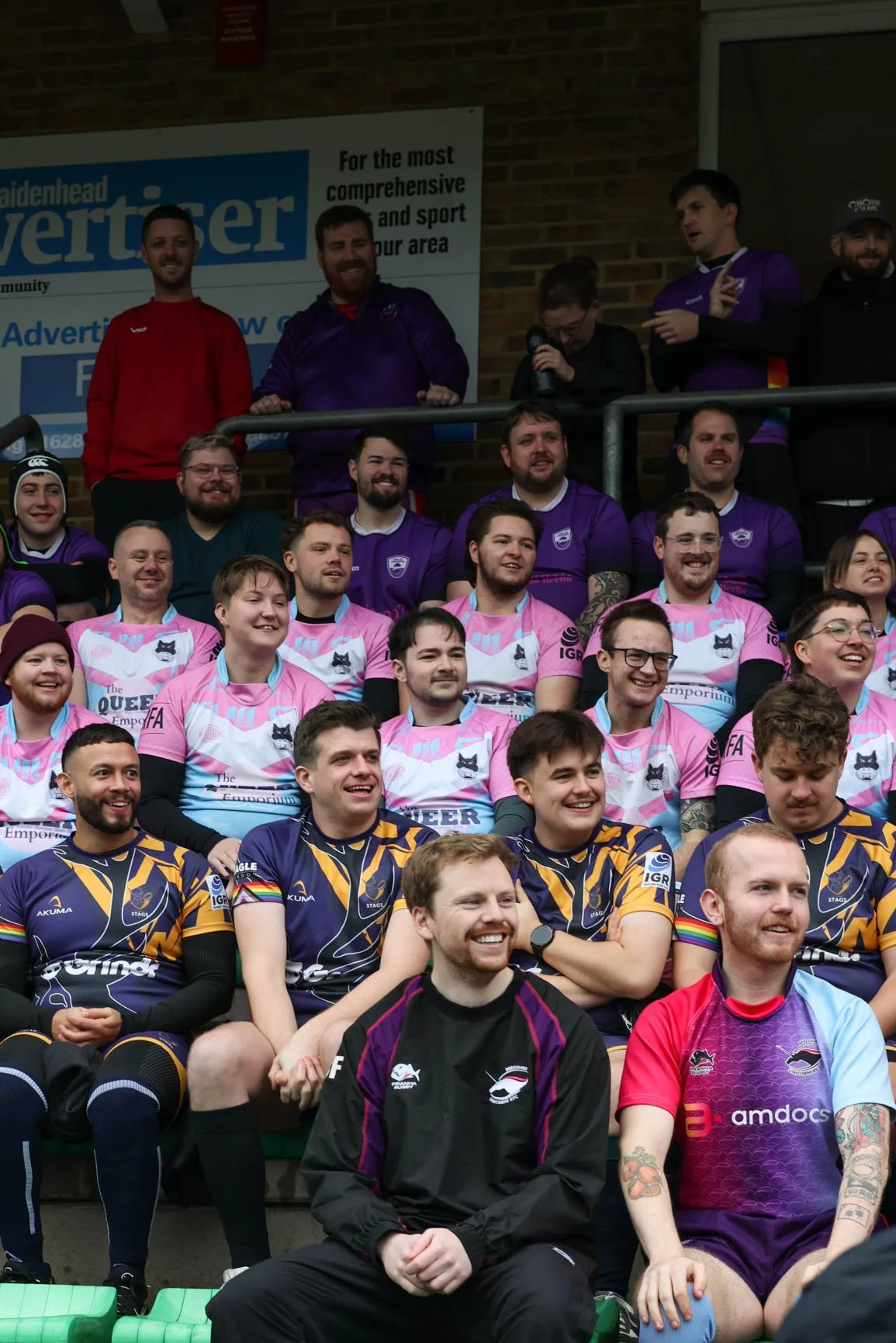 Group of rugby players and spectators, some in purple and pink jerseys, seated and standing on stadium bleachers.