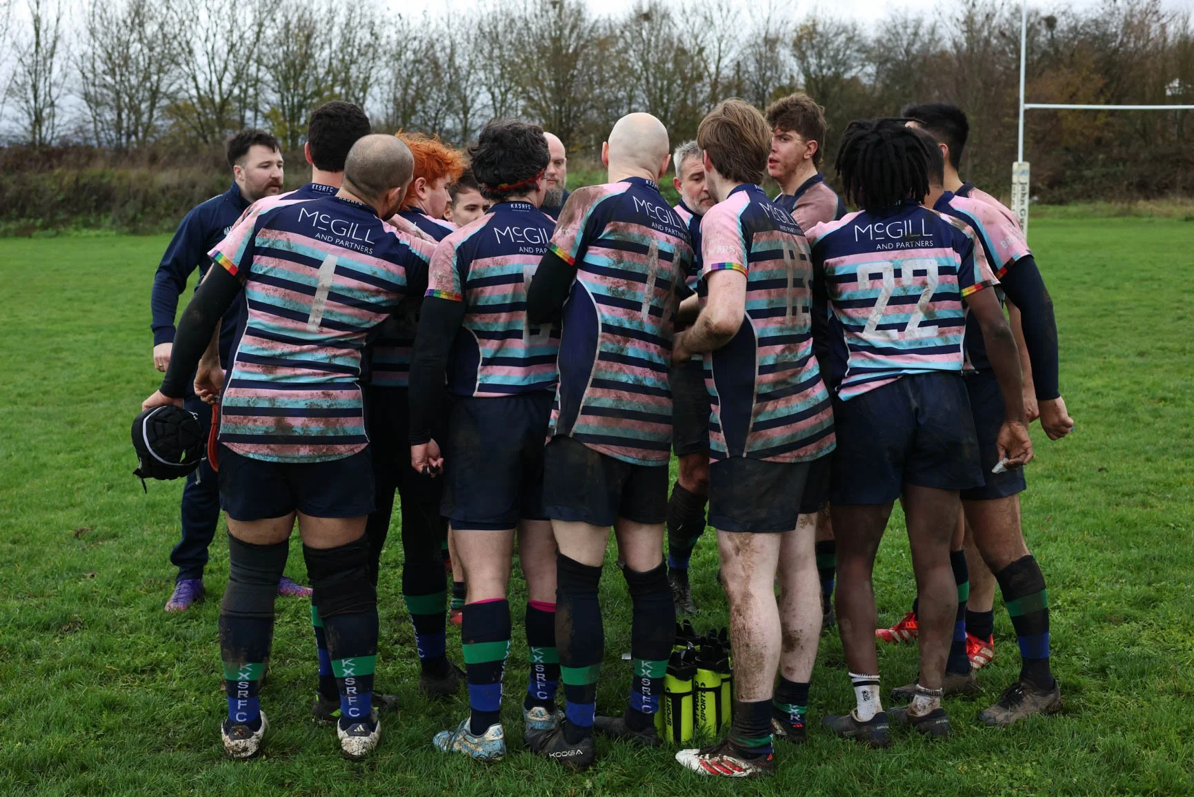 A group of rugby players huddled together on a grassy field during daytime, wearing matching uniforms with navy, pink, and green stripes, some holding a rugby ball and gear, with an overcast sky and a rugby goalpost in the background.
