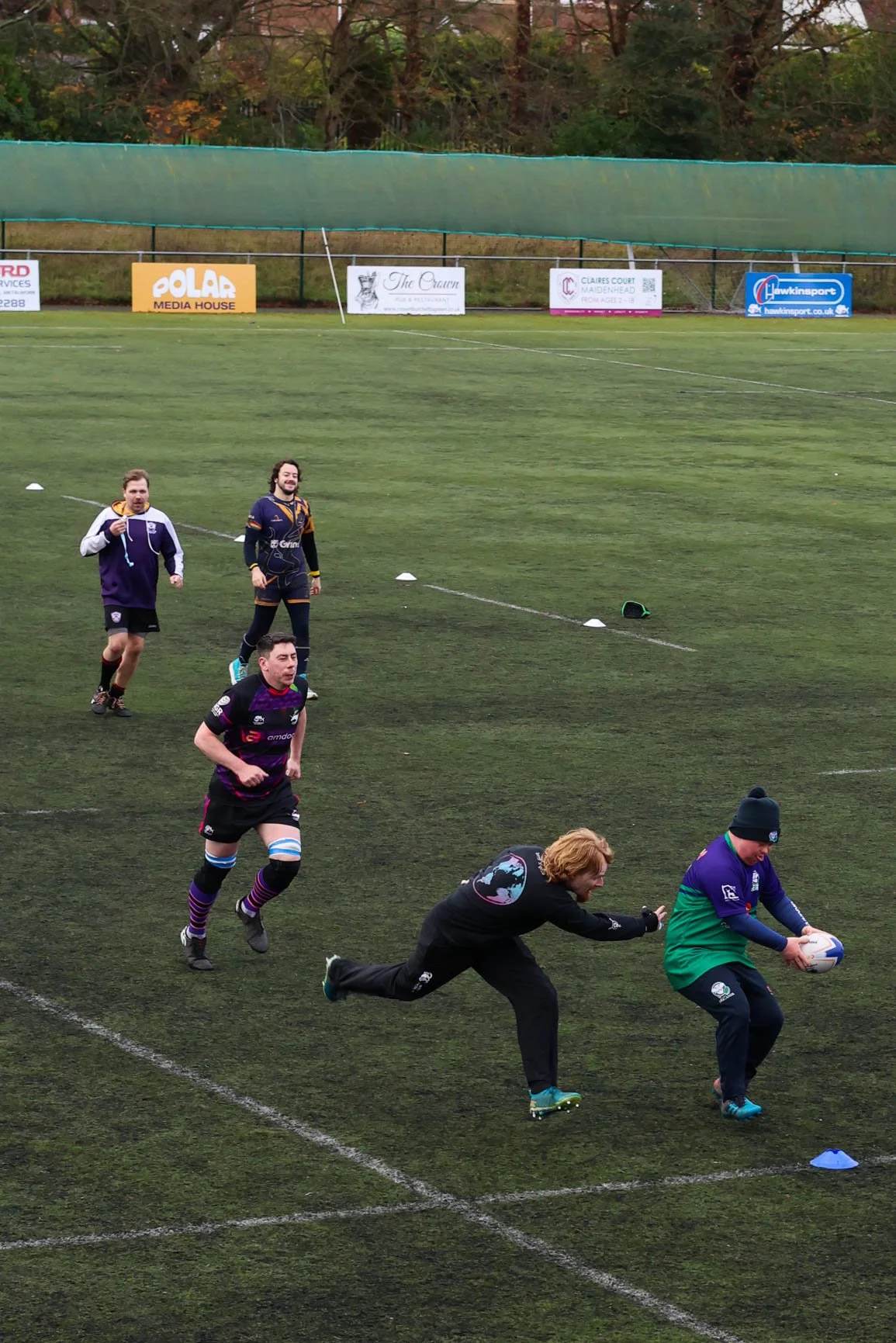 Rugby players practicing on an outdoor field, with one player running towards the ball, others running behind, and a coach or trainer in black reaching for the ball. The field has cones and advertisements in the background.