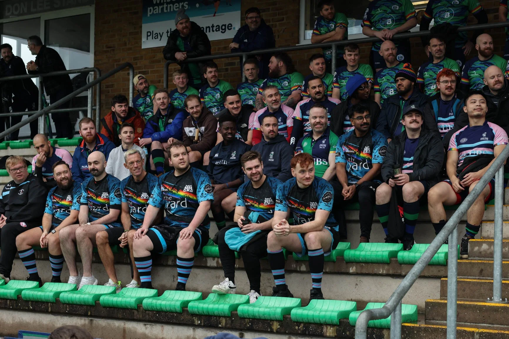 A group of rugby players and supporters sitting and standing on stadium bleachers, some wearing colorful team jerseys, others in casual clothing, with a brick wall and banners in the background.