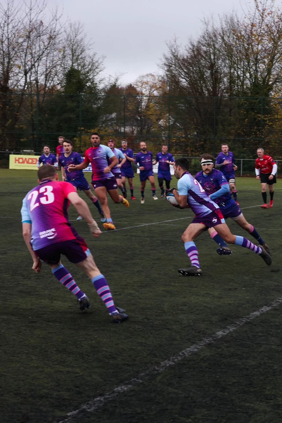 Rugby players in action on an outdoor field, with some players running and one holding the rugby ball, while others chase or defend. The scene takes place in overcast weather with leafless trees in the background.