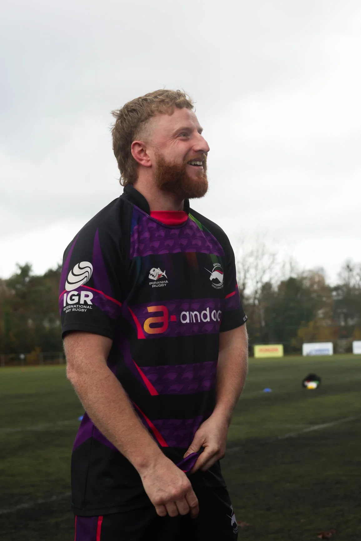 A man with red hair and a beard smiling and wearing a purple and black sports jersey.