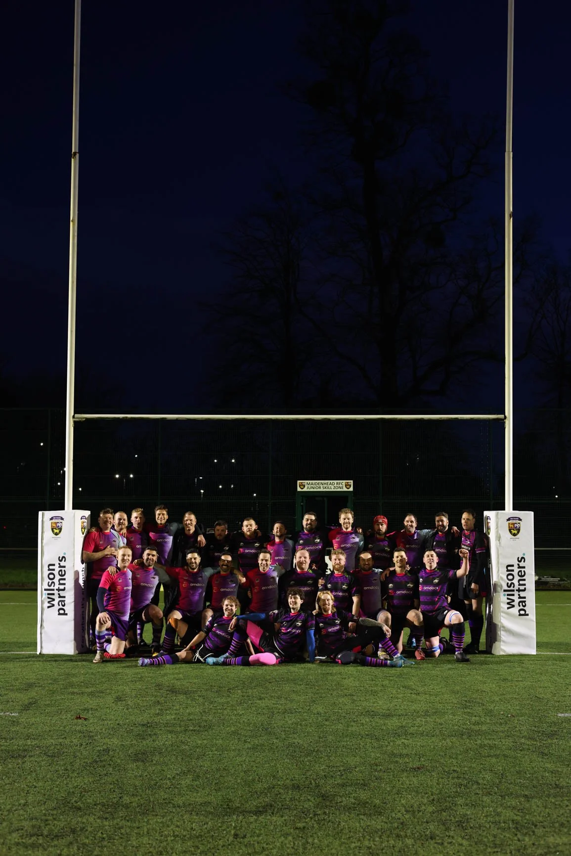 A rugby team in purple and pink uniforms posing together in front of a goalpost on a field at night, with trees and stadium lights in the background.