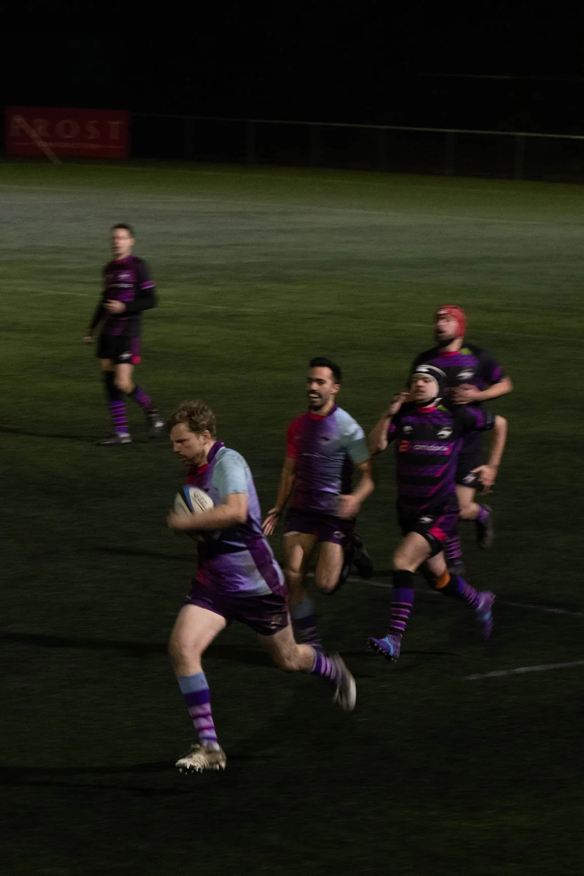 A group of rugby players running on a field at night during a match, dressed in team uniforms with a rugby ball in one player's hands.