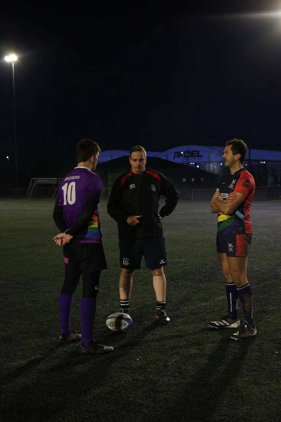 Three men on a rugby field at night, two wearing colorful rugby uniforms and one in black sportswear, standing near a rugby ball.