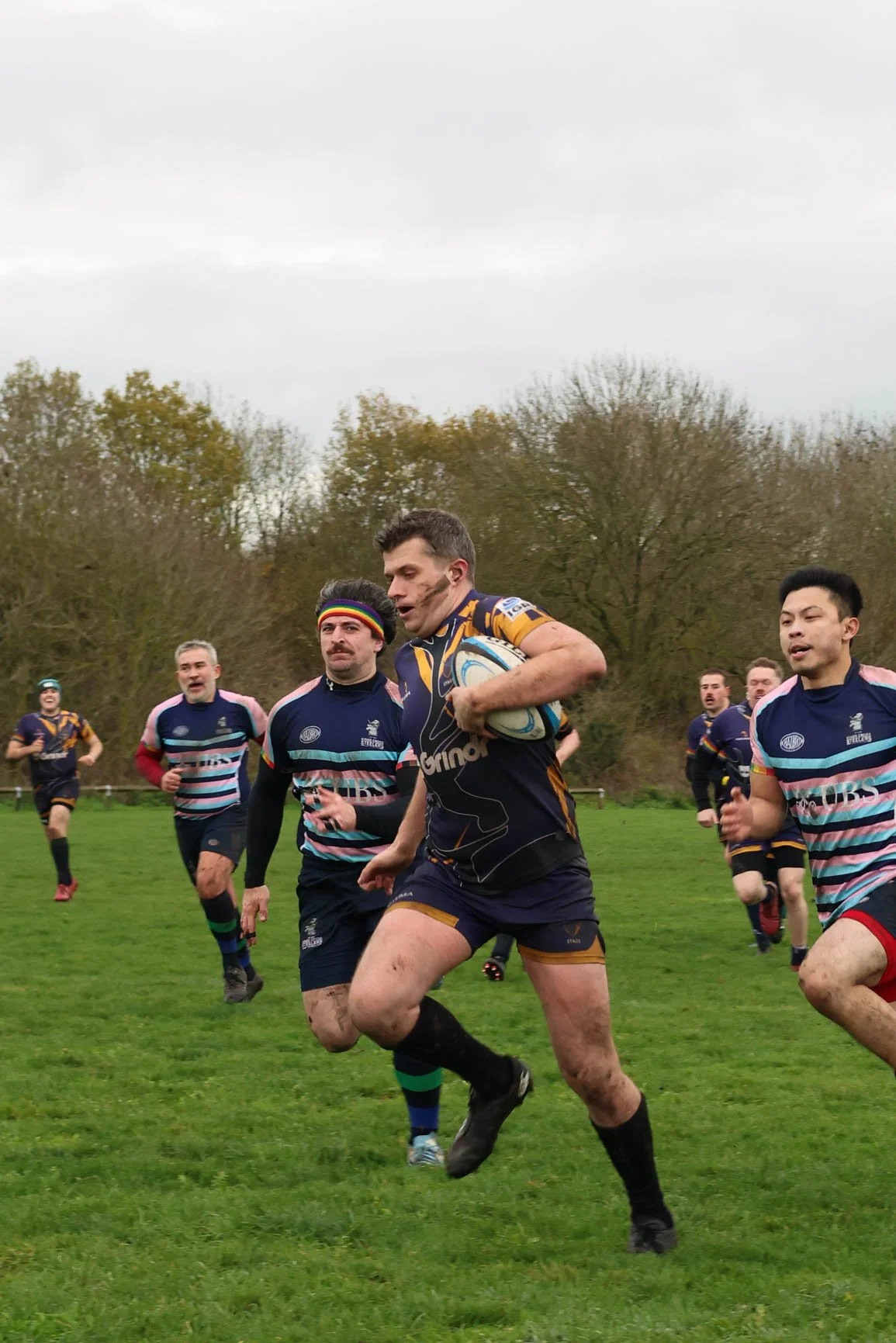 Rugby players running on a grassy field during a game, with trees and overcast sky in the background.
