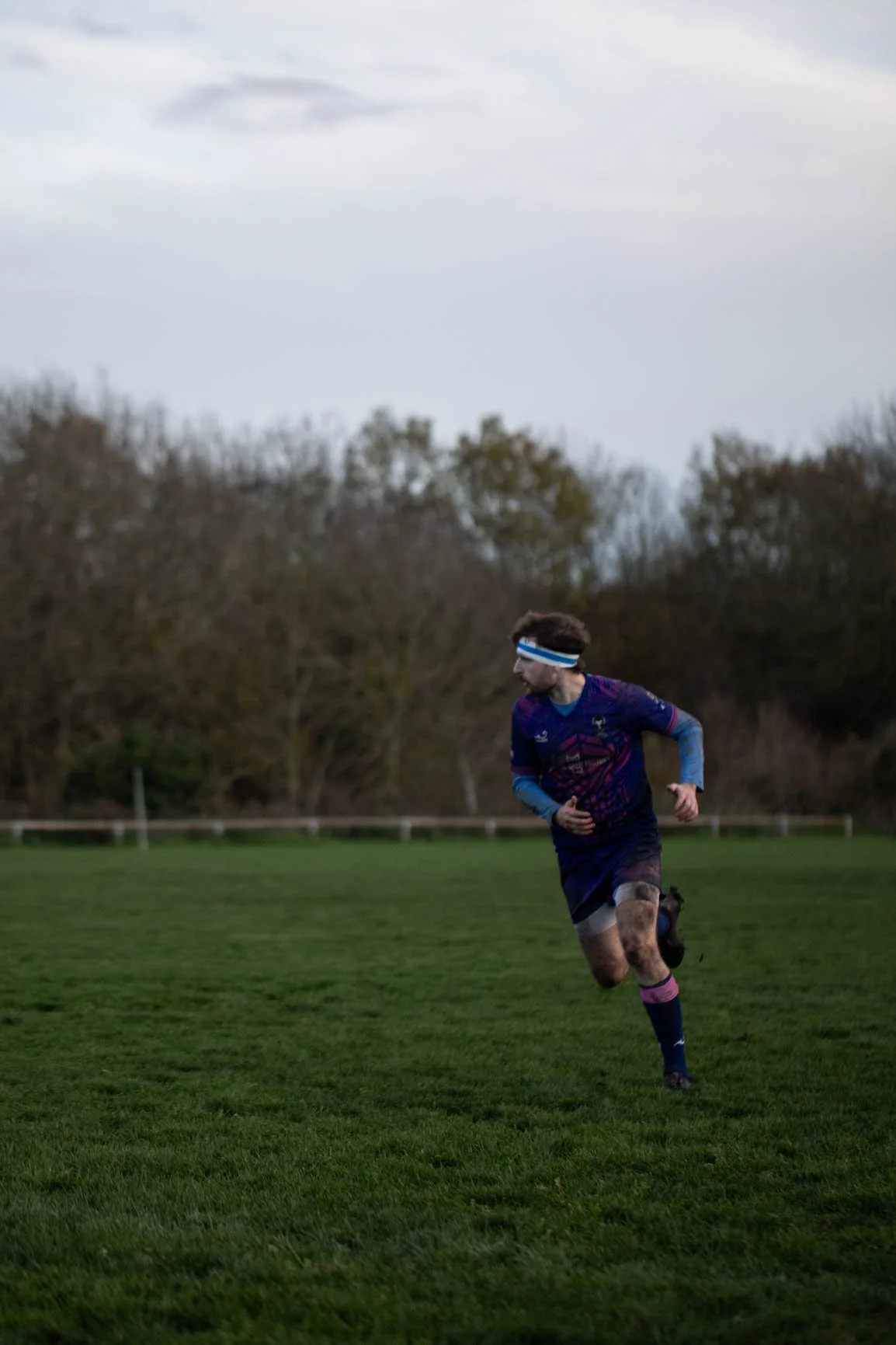 A rugby player running on a grassy field, wearing a blue and purple uniform with a headband, during daytime with cloudy sky.