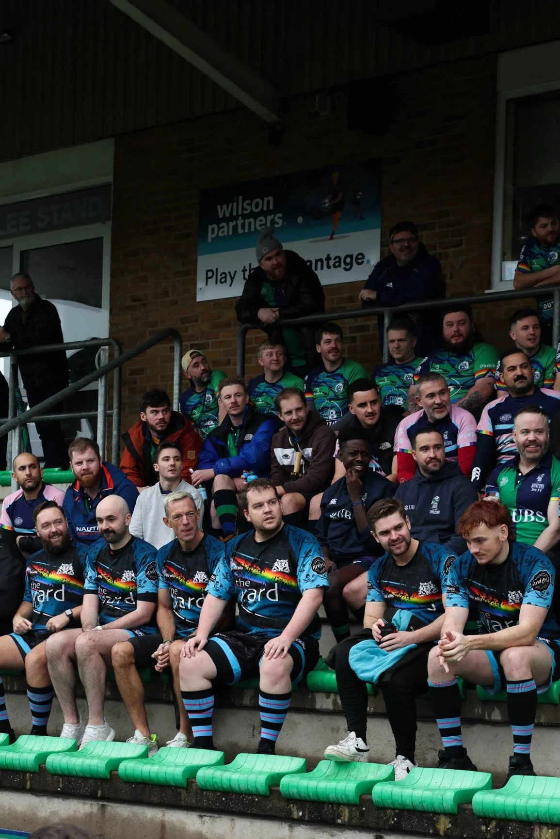 Group of rugby players and spectators sitting in stadium stands, wearing colorful rugby jerseys and casual clothing, with a banner in the background promoting Wilson Partners.