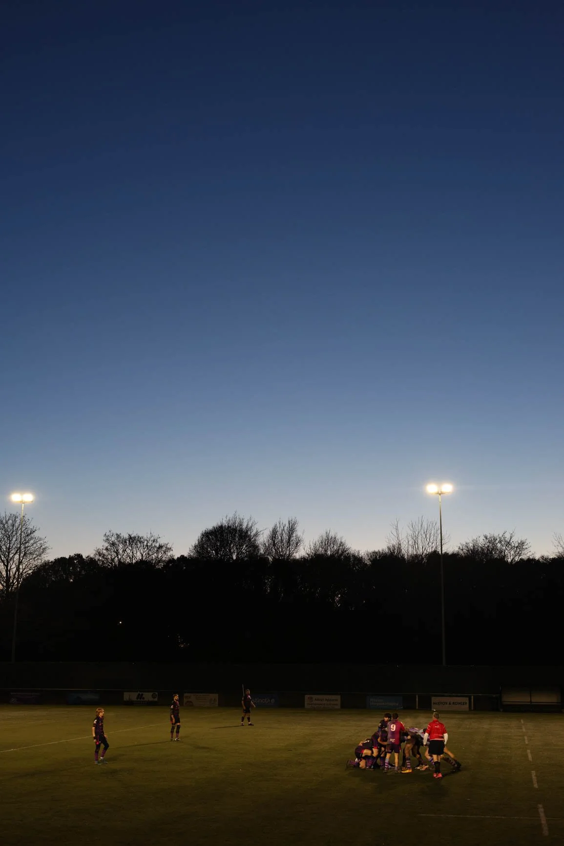 A rugby match taking place on a field at dusk with floodlights and a dark sky, and players huddled together.