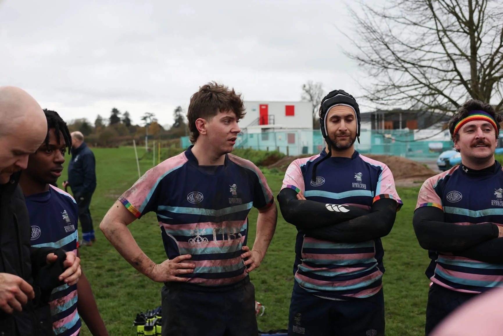 Group of rugby players in rugby uniforms standing outdoors on a cloudy day, listening to a coach.