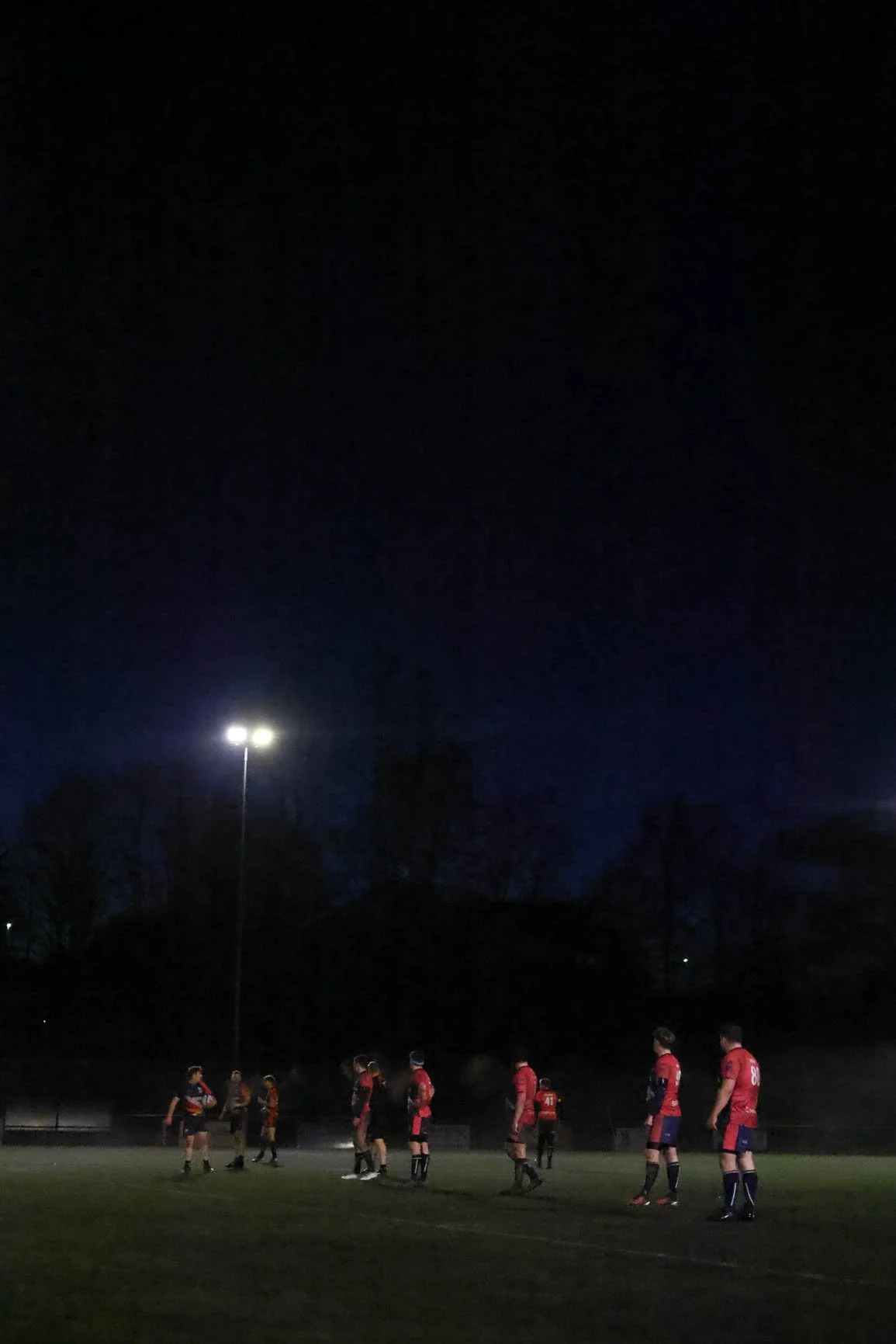 A group of soccer players in red uniforms standing on a field at night under a dark sky with a bright stadium light.