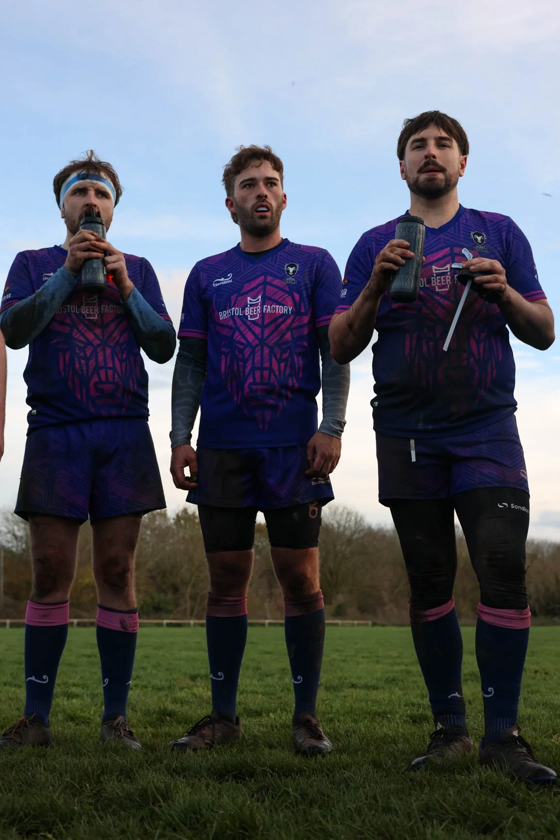 Three male rugby players wearing purple jerseys standing on a grassy field, holding water bottles, with a light blue sky and trees in the background.