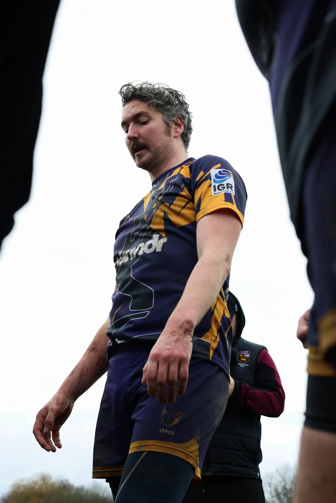 A male rugby player on the field wearing a navy blue and yellow uniform, surrounded by teammates, with a cloudy sky in the background.