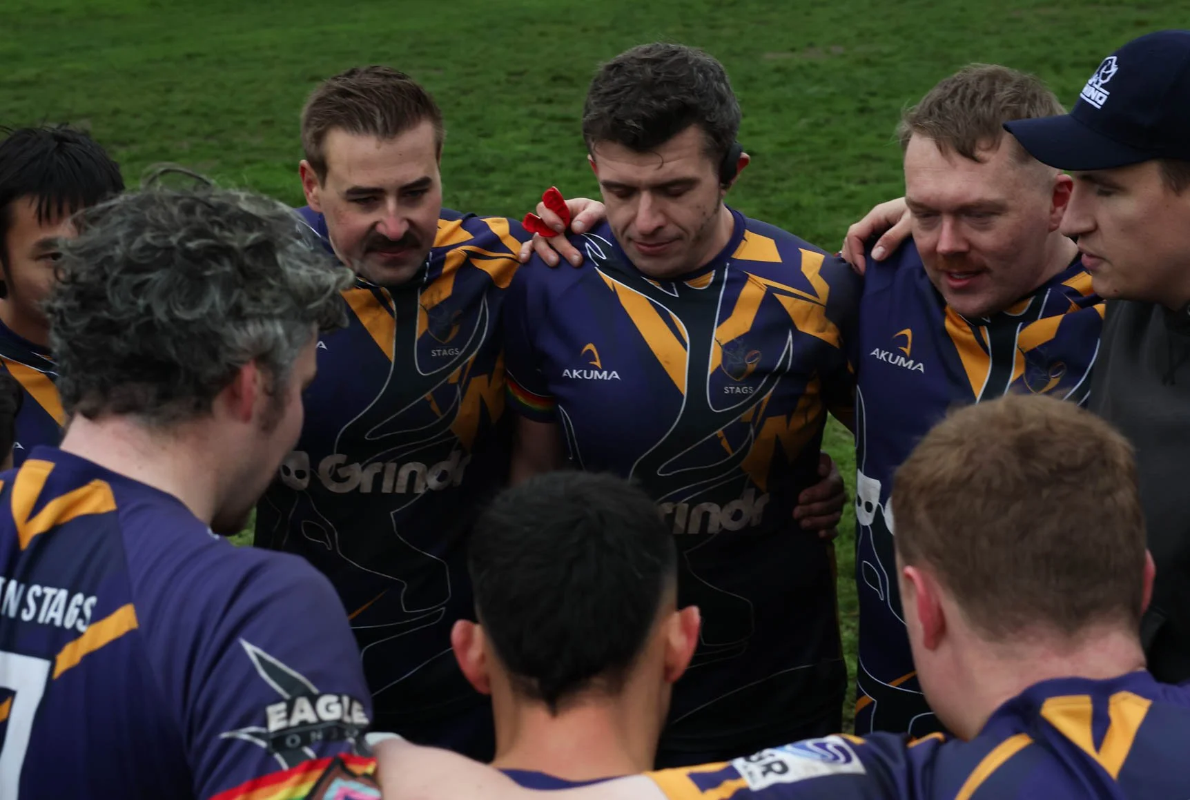 A group of rugby players in dark blue and yellow jerseys huddled together in a team pre-game pep talk on the field.
