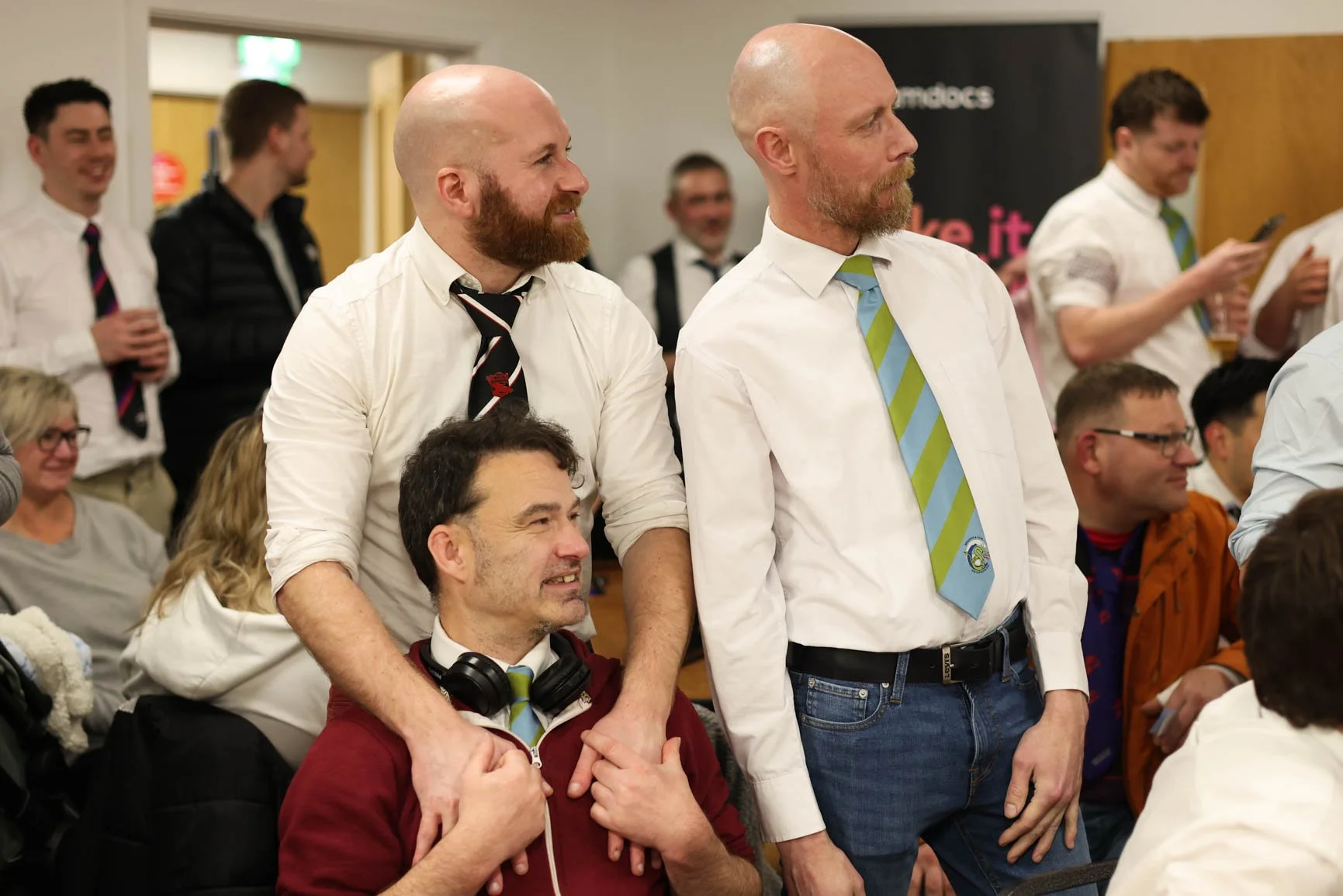 A group of people at an indoor event, some standing and some sitting, smiling and engaging with each other. Two men with bald heads and beards are prominently standing in the foreground, holding hands with a man seated in front of them. The scene app