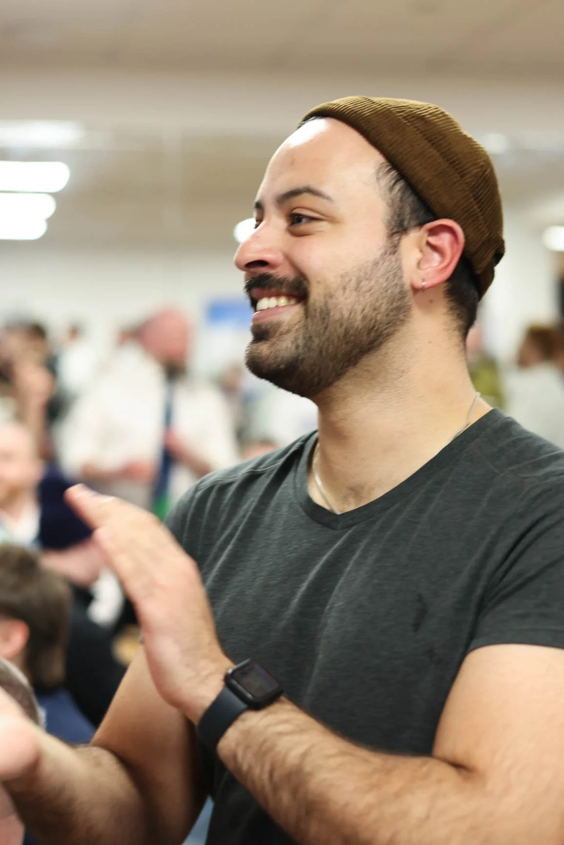 A smiling man with a beard and mustache, wearing a brown knit cap and black t-shirt, standing in a crowded indoor setting.