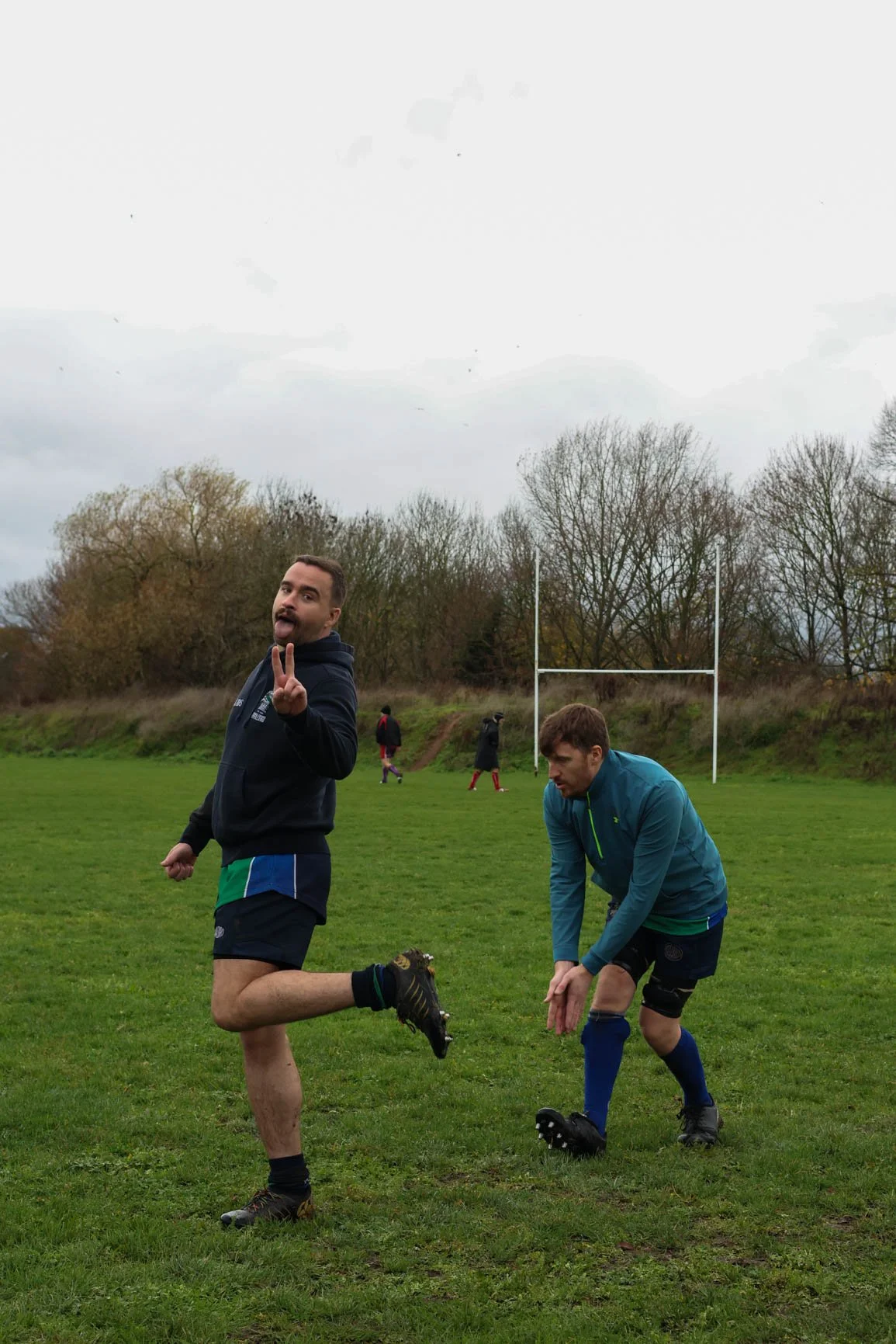 Two men dressed in sportswear playing rugby on a grassy field with goal posts, trees, and cloudy sky in the background. One man is making a peace sign and sticking out his tongue, while the other is bending down, possibly tying his shoe.