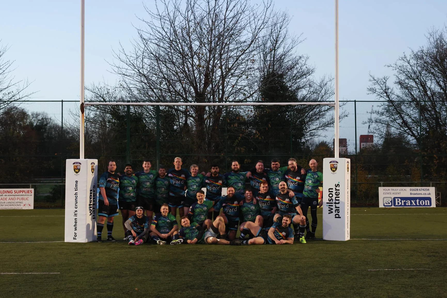 A group of rugby players in colorful uniforms posing together on a rugby field near goalposts. They are smiling and standing in front of a fence with trees and advertising signs in the background.