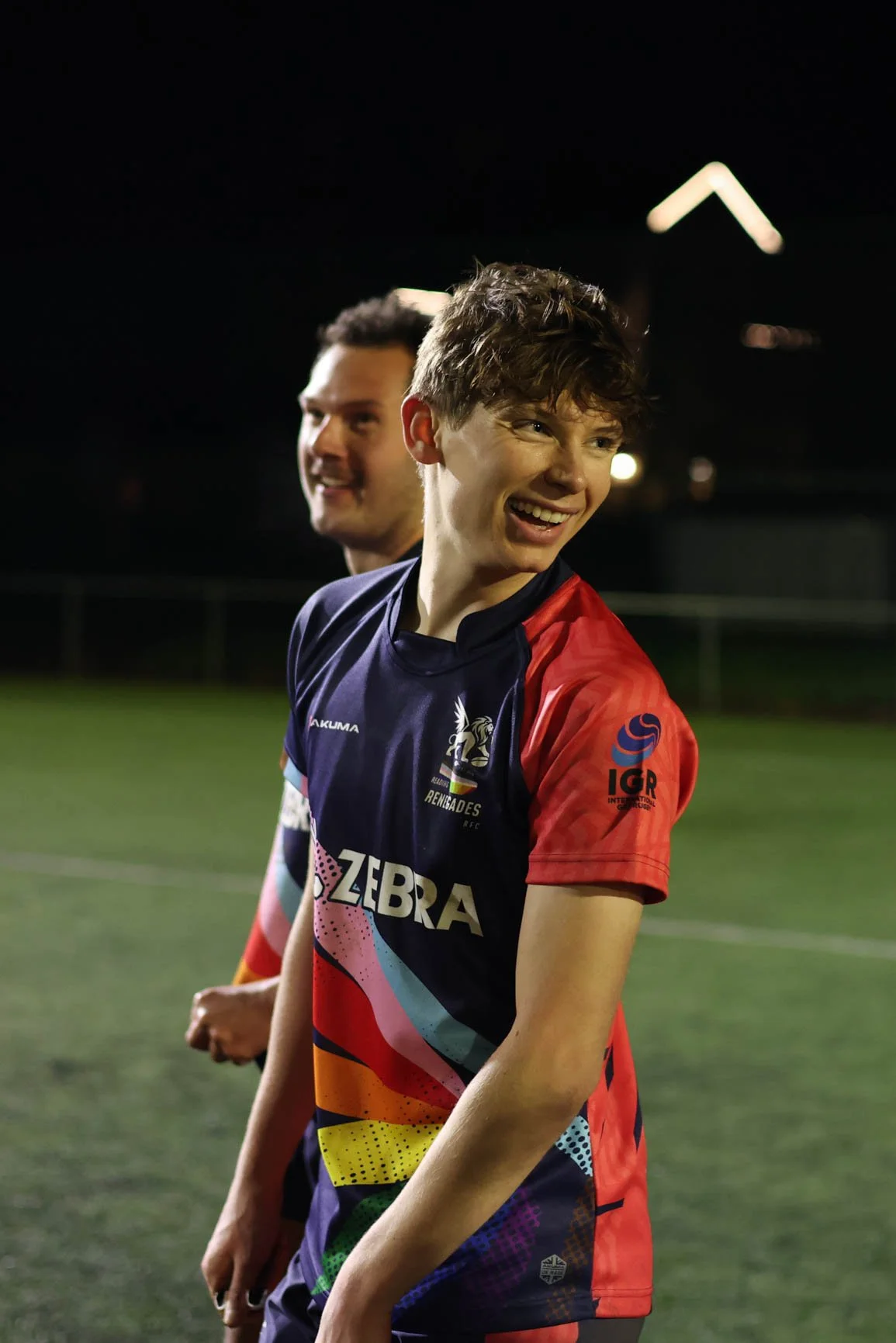 Two young male rugby players stand on a field at night, smiling and wearing colorful jerseys with sponsor logos. The player in the foreground is smiling with curly hair, while the other player is in the background with short dark hair.