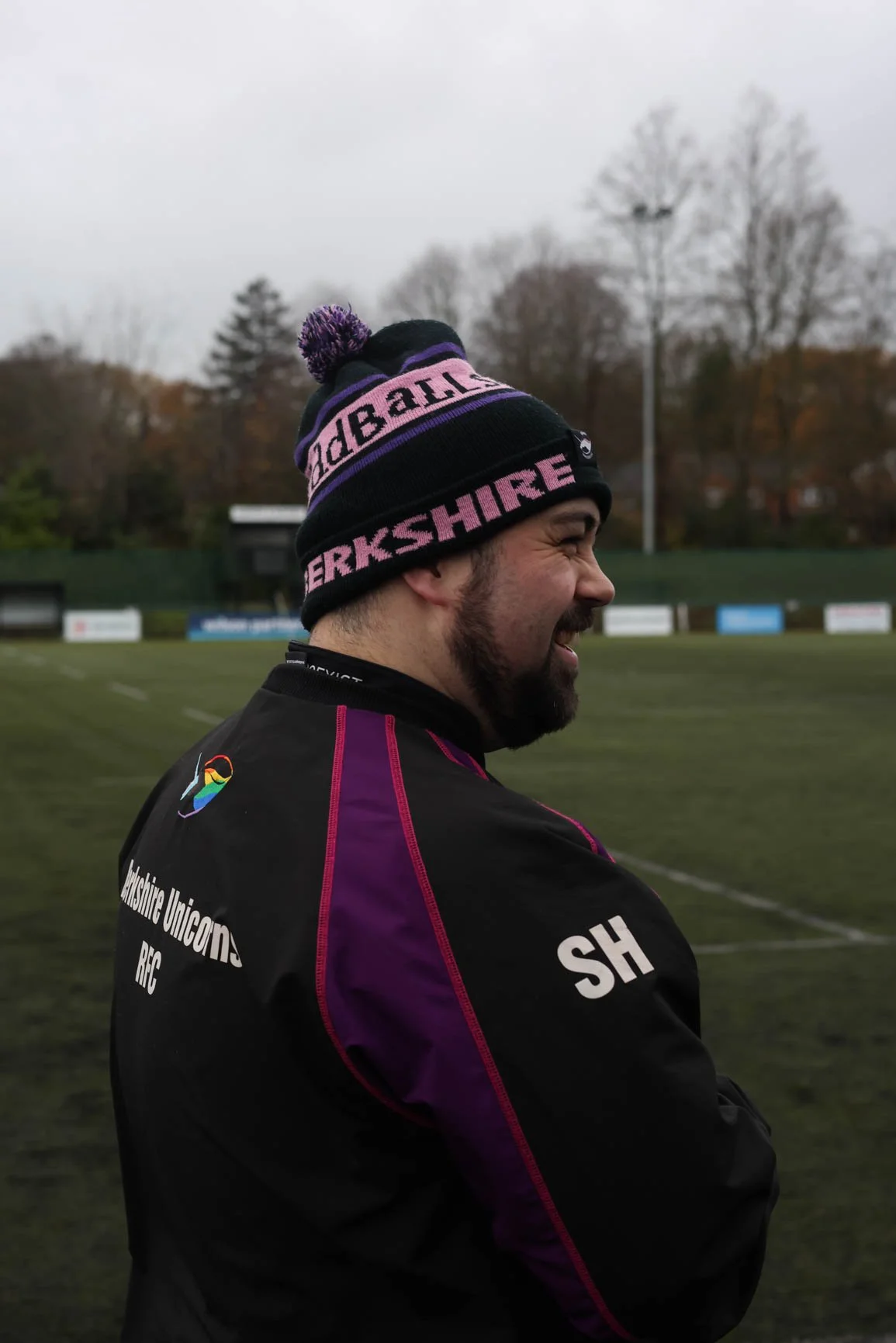 A man smiling in profile wearing a Warwickshire rugby jacket and a black, purple, and pink beanie hat with a pom-pom on top, standing on a rugby field with trees and a cloudy sky in the background.