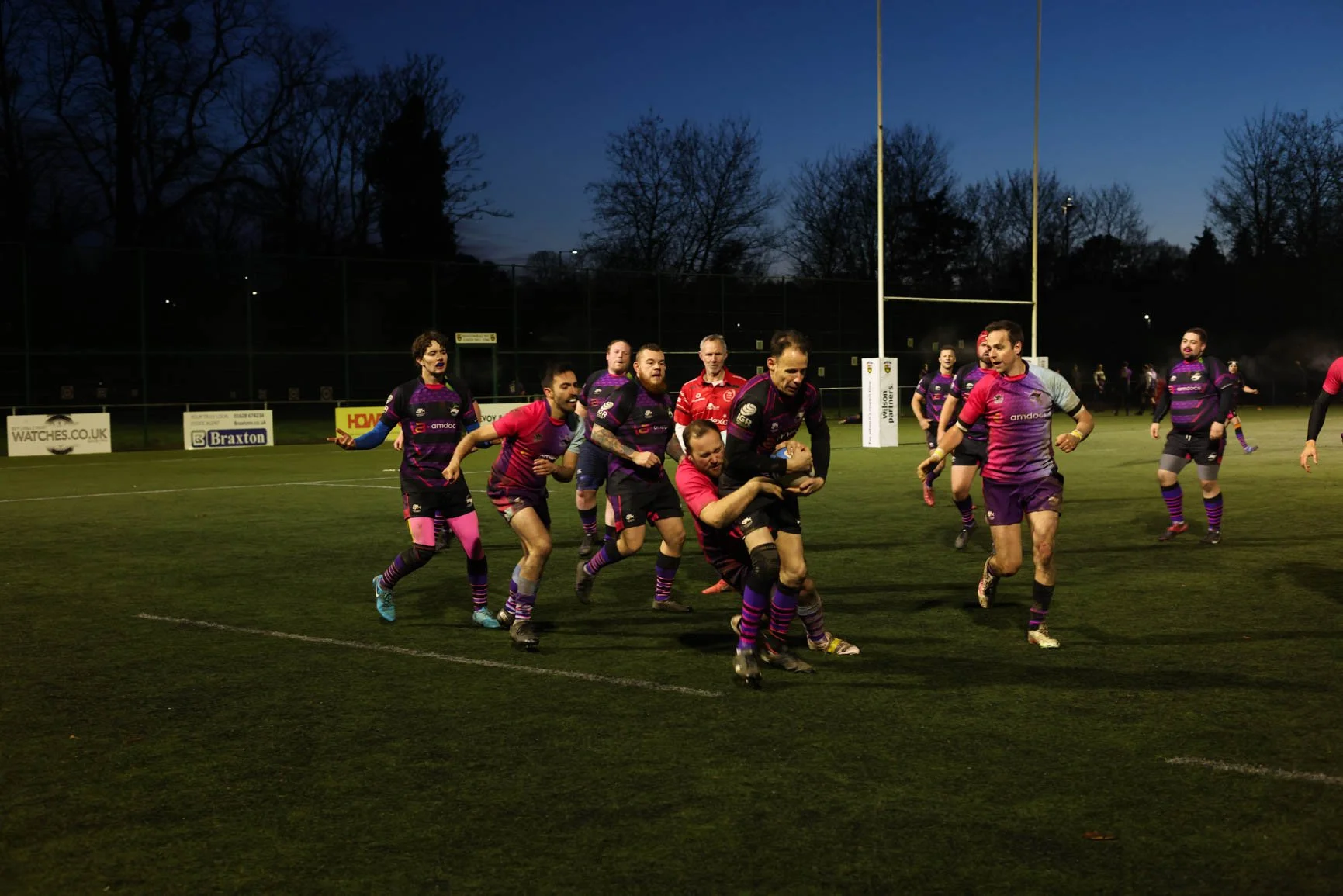 Rugby players in purple, pink, and black uniforms celebrating on a rugby field during dusk, with a goalpost and trees in the background.