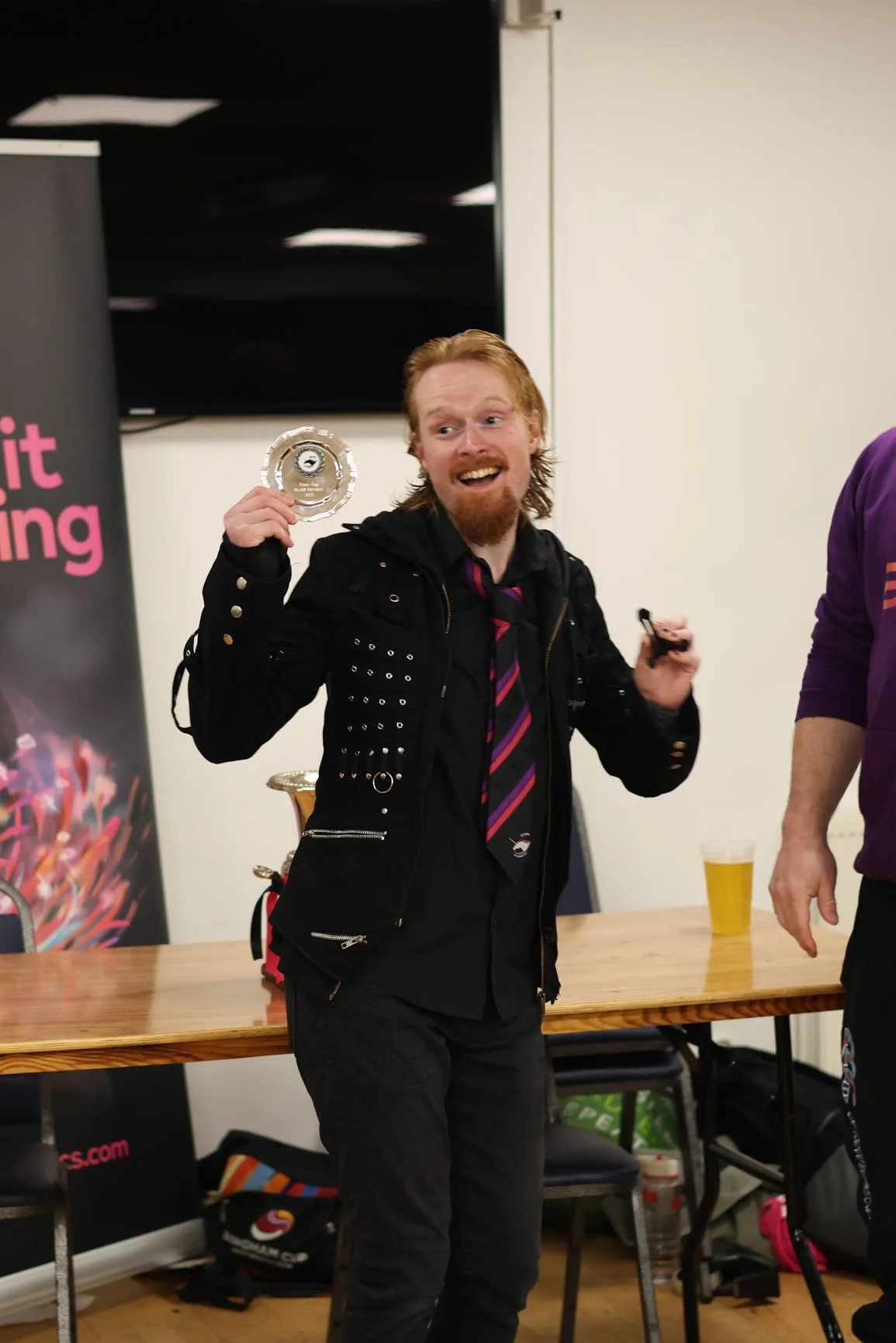 A man with long red hair and a beard, wearing a black jacket with metal studs and a black shirt with a purple and pink striped tie, holding a clear glass award in his right hand and smiling at a celebration or event.