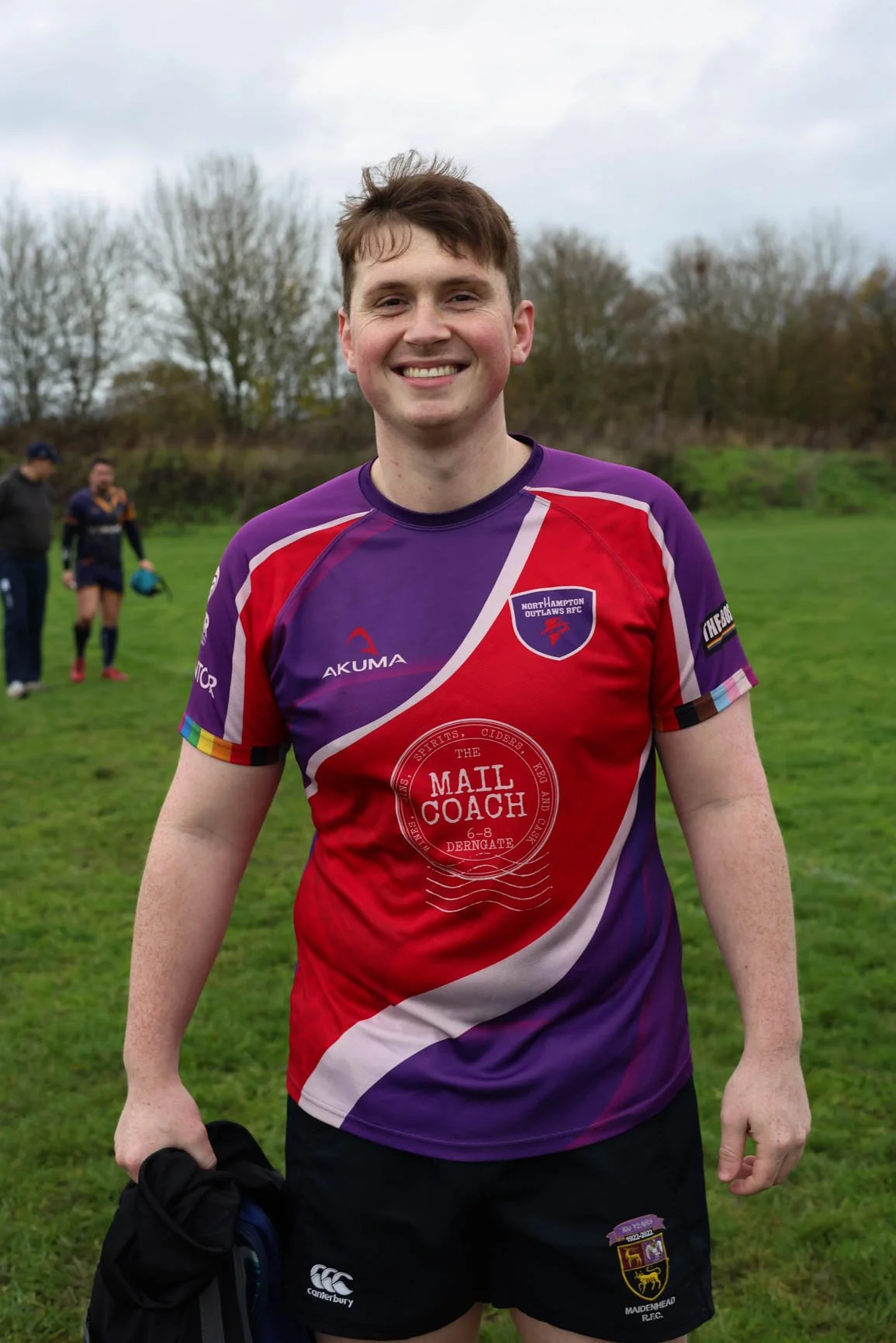 A young man smiling outdoors in sports attire, holding a jacket, with a grassy field and other people in the background.