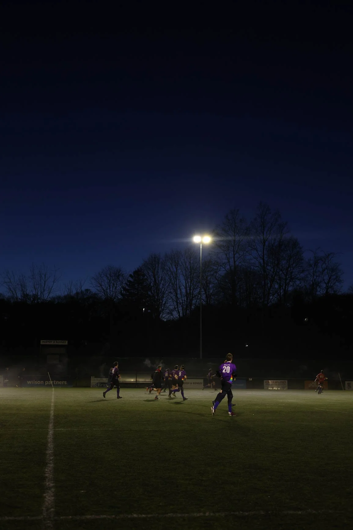 Football players in purple jerseys running on the field during a game at night under stadium lights, with a dark sky and trees in the background.