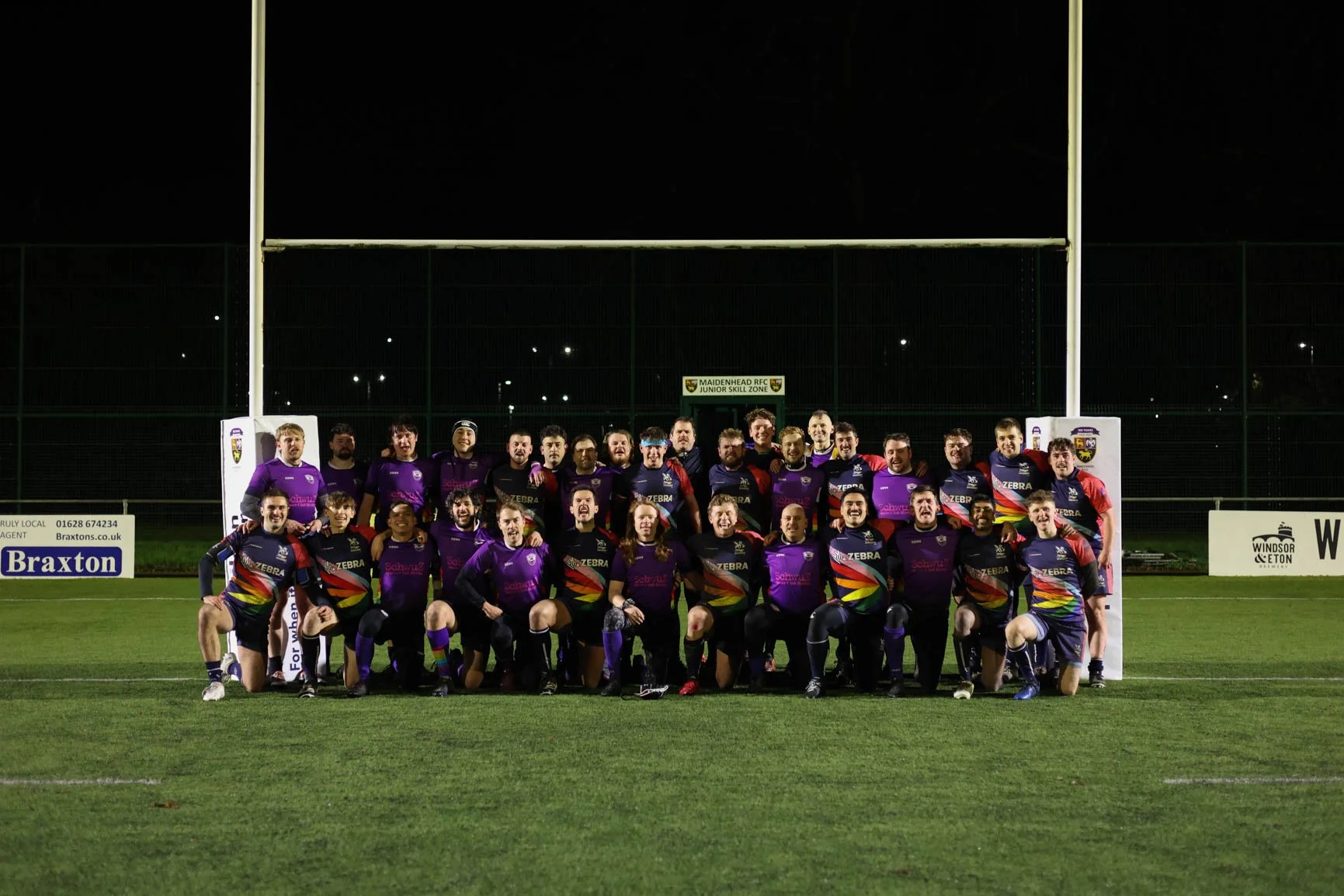 A group of rugby players in purple and colorful uniforms posing on a rugby field at night with goalposts in the background.