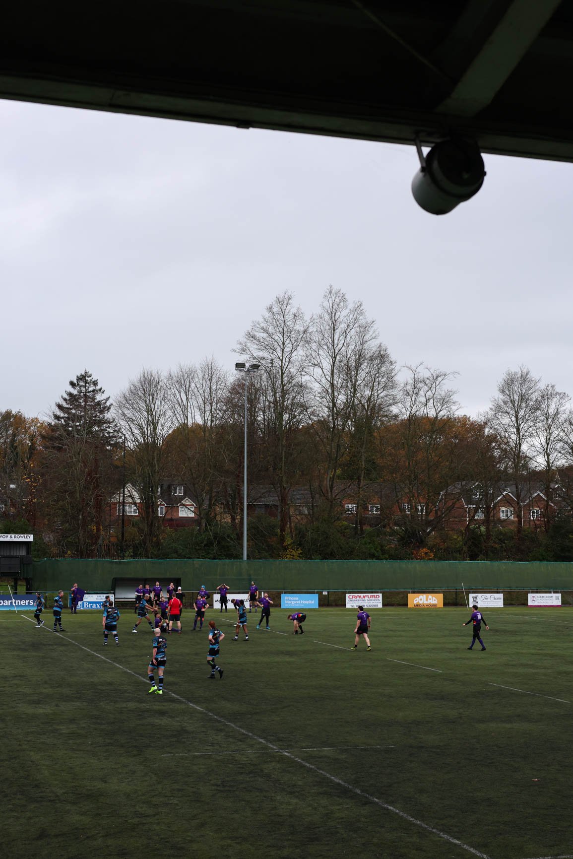 A rugby match underway on a field surrounded by trees, with players in purple and blue uniforms. There are advertisements along the fence, and leafless trees and residential houses in the background. The sky is overcast.