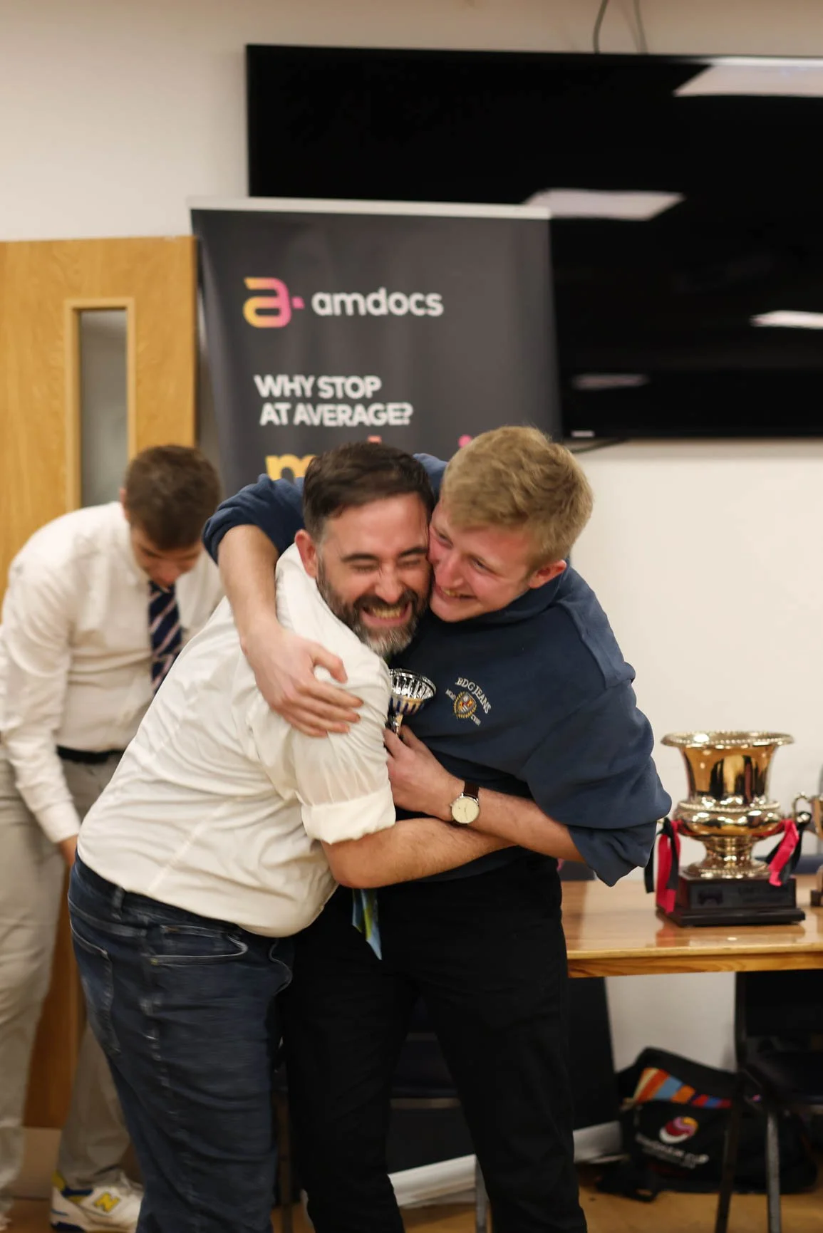 Two men hugging and celebrating, one holding a trophy, at a social event with a banner in the background and a table with a trophy on it.