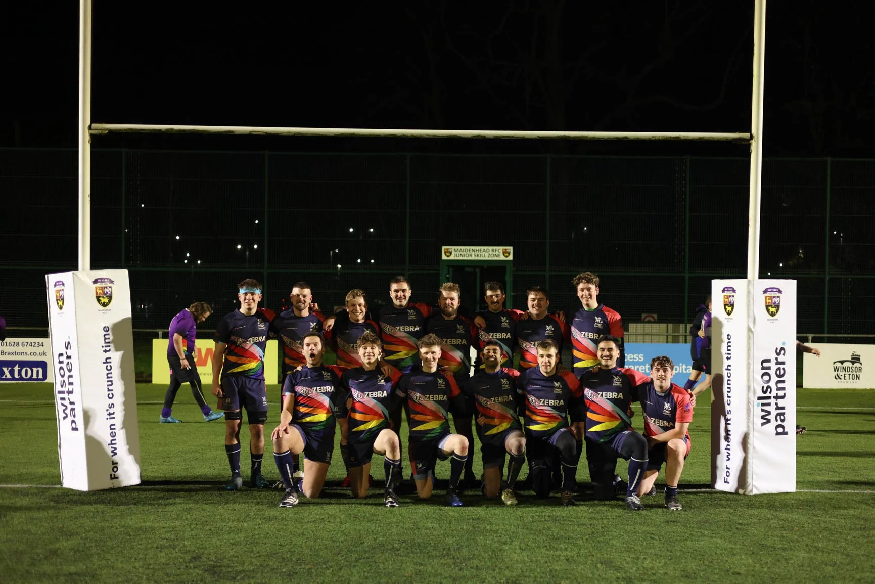 Group of rugby players in colorful team jerseys posing for a photo on a rugby field at night, with goalposts and advertising banners in the background.