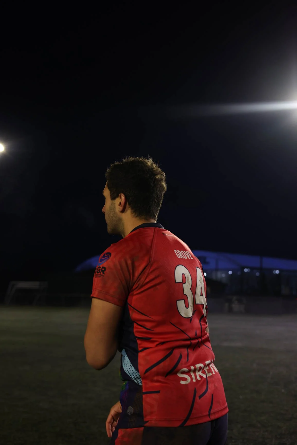 A male athlete wearing a red sports jersey with the number 34 and the name 'GROVE' on the back, standing on a sports field at night, illuminated by stadium lights, with a dark sky in the background.